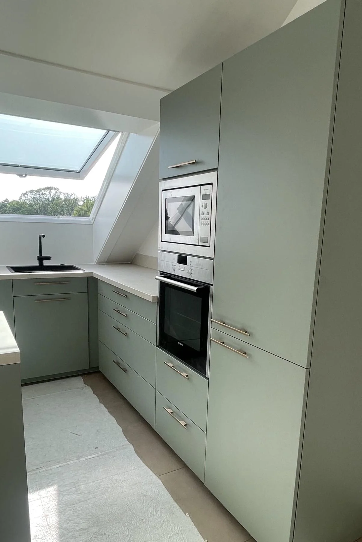 Cuisine moderne avec armoires vertes, four, micro-ondes, évier sous fenêtre inclinée, vue sur le ciel et la nature.