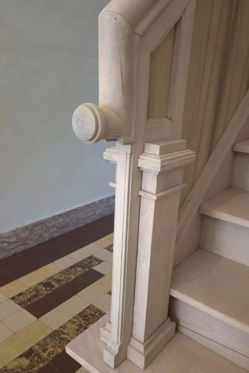 Détail d'une balustrade en bois non peinte avec nouvelleiton de profil classique, posée le long d'un escalier en bois massif. Le sol en carrelage présente un motif de carreaux beige et marron.