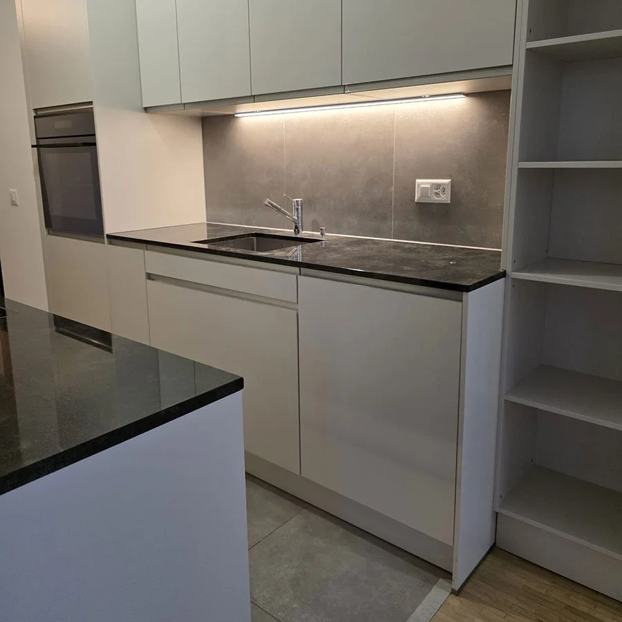 Cuisine moderne avec un évier, des armoires blanches, des comptoirs en pierre noire, une prise électrique sur le mur et une étagère vide.