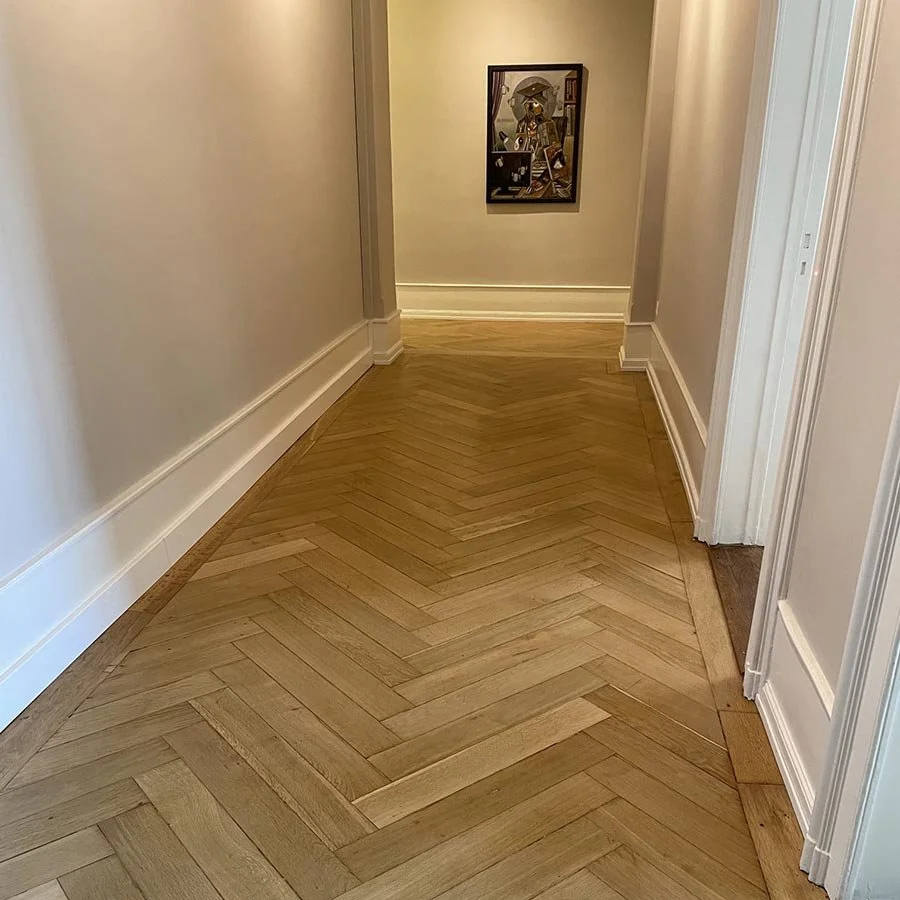 Un couloir intérieur avec un sol en parquet en chevrons, des murs clairs et une peinture encadrée au fond.