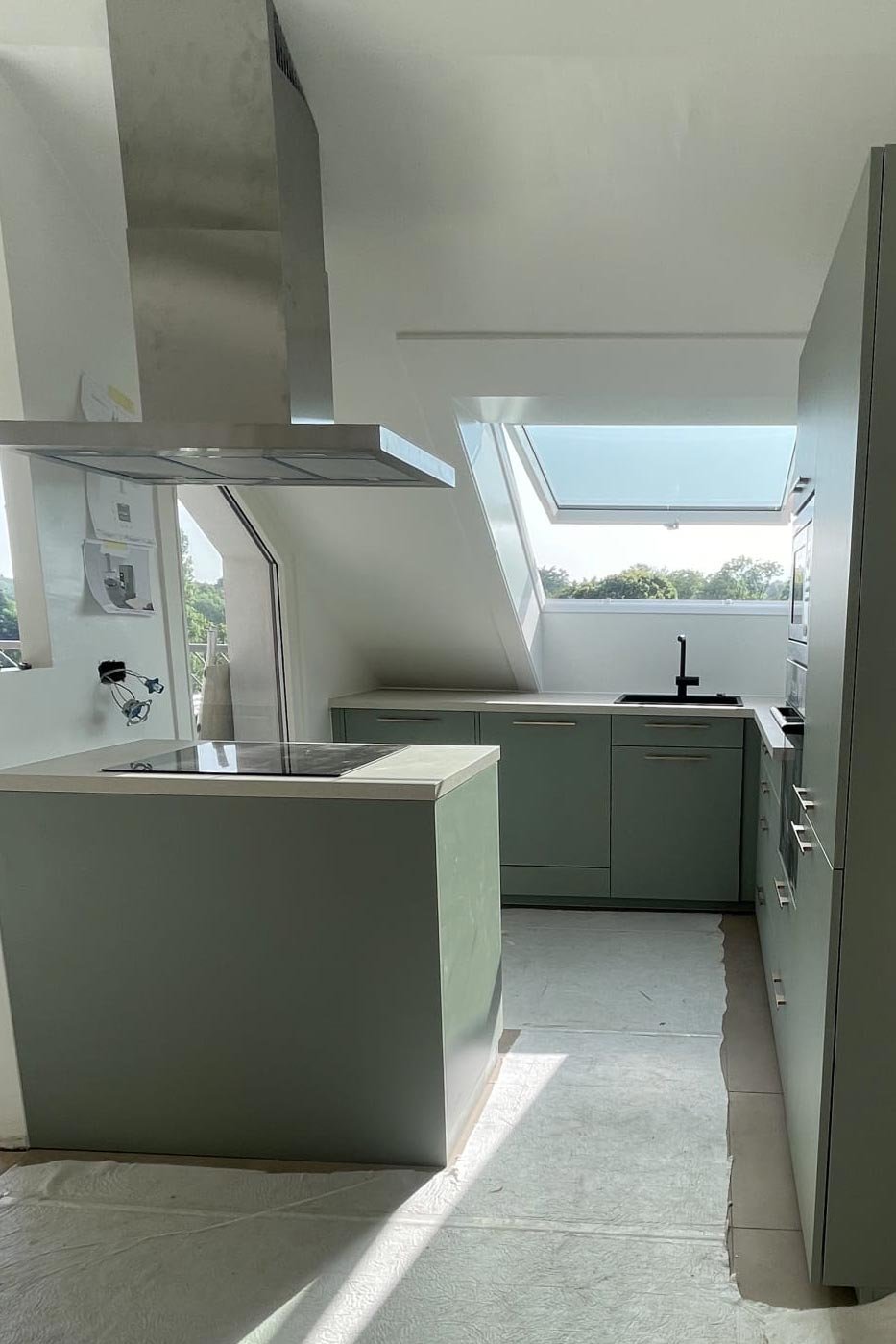 Cuisine moderne avec fenêtres en angle, plan de travail vert pastel, évier noir, hotte en acier inoxydable, et sol en béton brut.