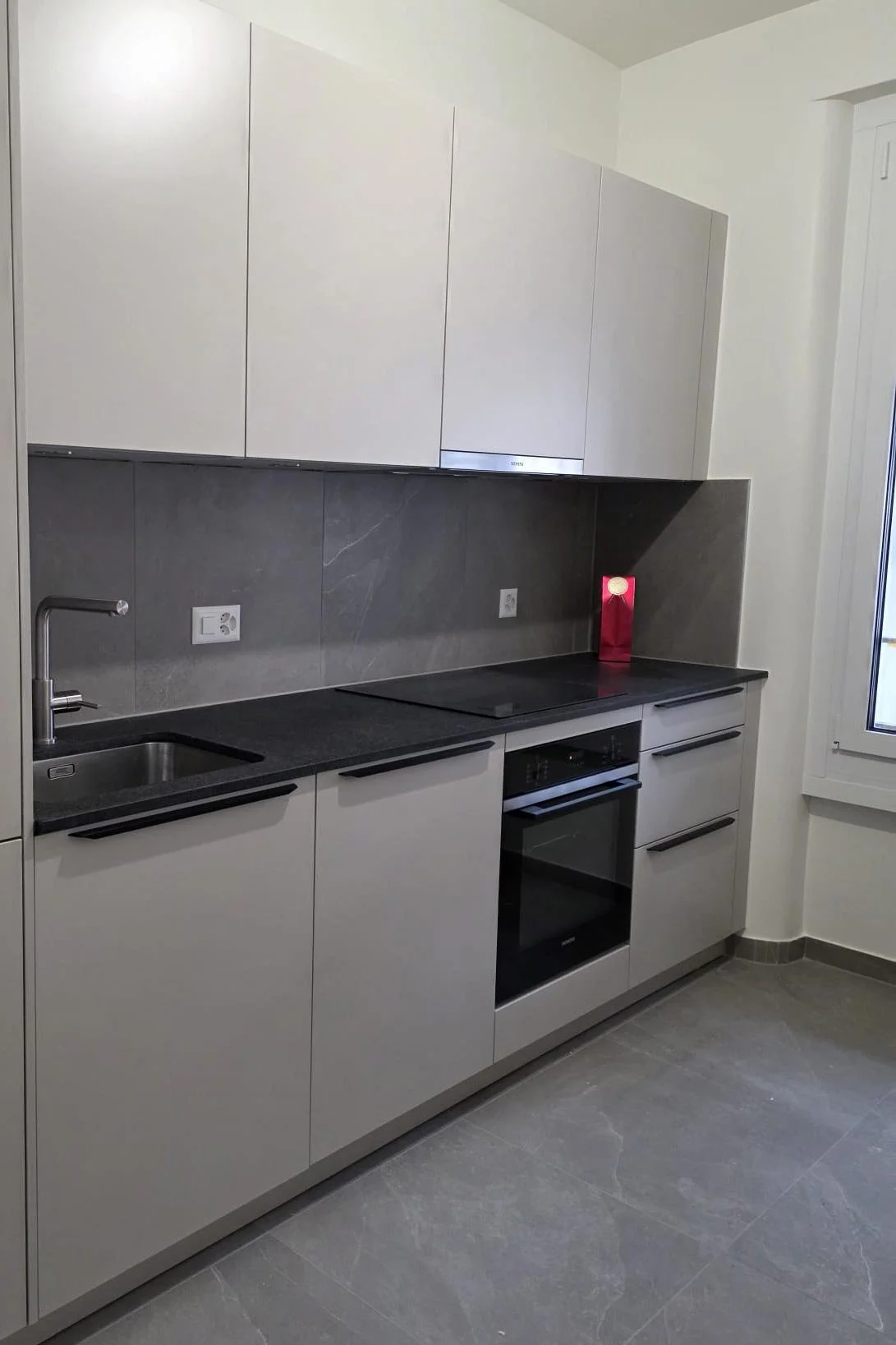 Cuisine moderne avec une évier, un four intégré, des armoires blanches et une plaque de cuisson noire, avec un mur en carrelage gris foncé.