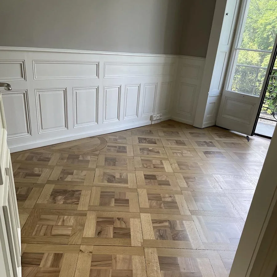 Pièce lumineuse avec parquet en bois et murs ornés de lambris blancs, grande fenêtre donnant sur un balcon avec vue sur la verdure.