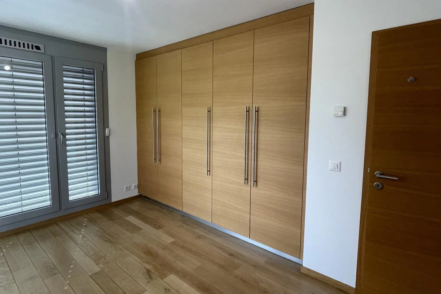 Une pièce intérieure avec un grand placard en bois clair, une porte en bois et une porte-fenêtre avec volets roulants métalliques.