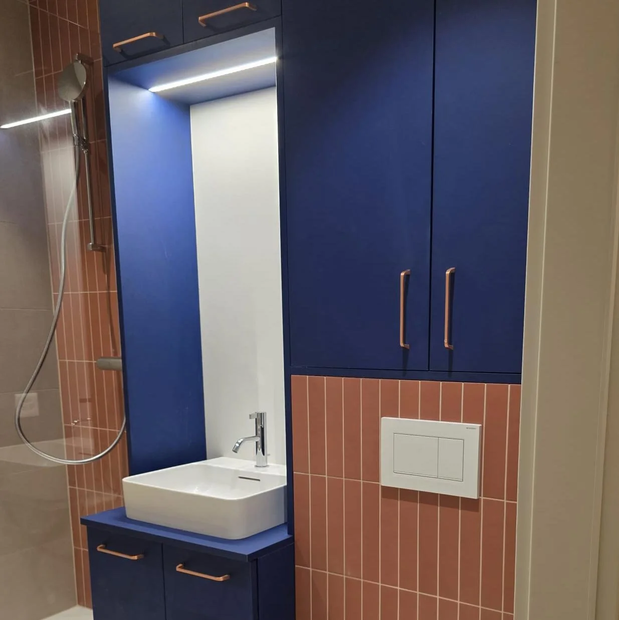 Salle de bain avec un miroir, un lavabo blanc, des armoires bleues avec des poignées en cuivre, carrelage rose au mur.