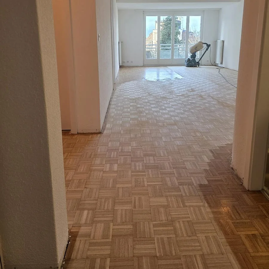 Une pièce vide avec un sol en parquet en cours de rénovation, un aspirateur industriel dans le fond, avec des portes-fenêtres donnant sur un balcon et des bâtiments environnants