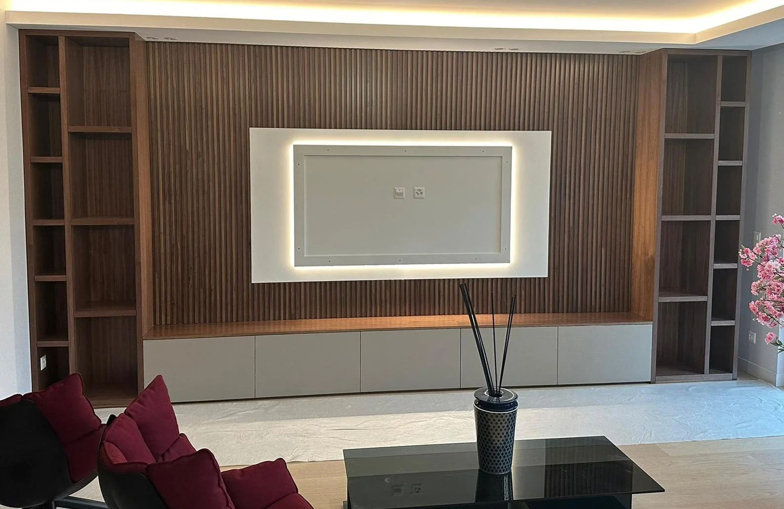 Mur en bois avec étagères encadrant un espace pour une télévision, éclairé par un éclairage intégré. Un meuble bas gris en dessous, une table en verre avec un vase contenant des bâtons d'encens, et un canapé rouge avec des coussins. Une fleur de ceri