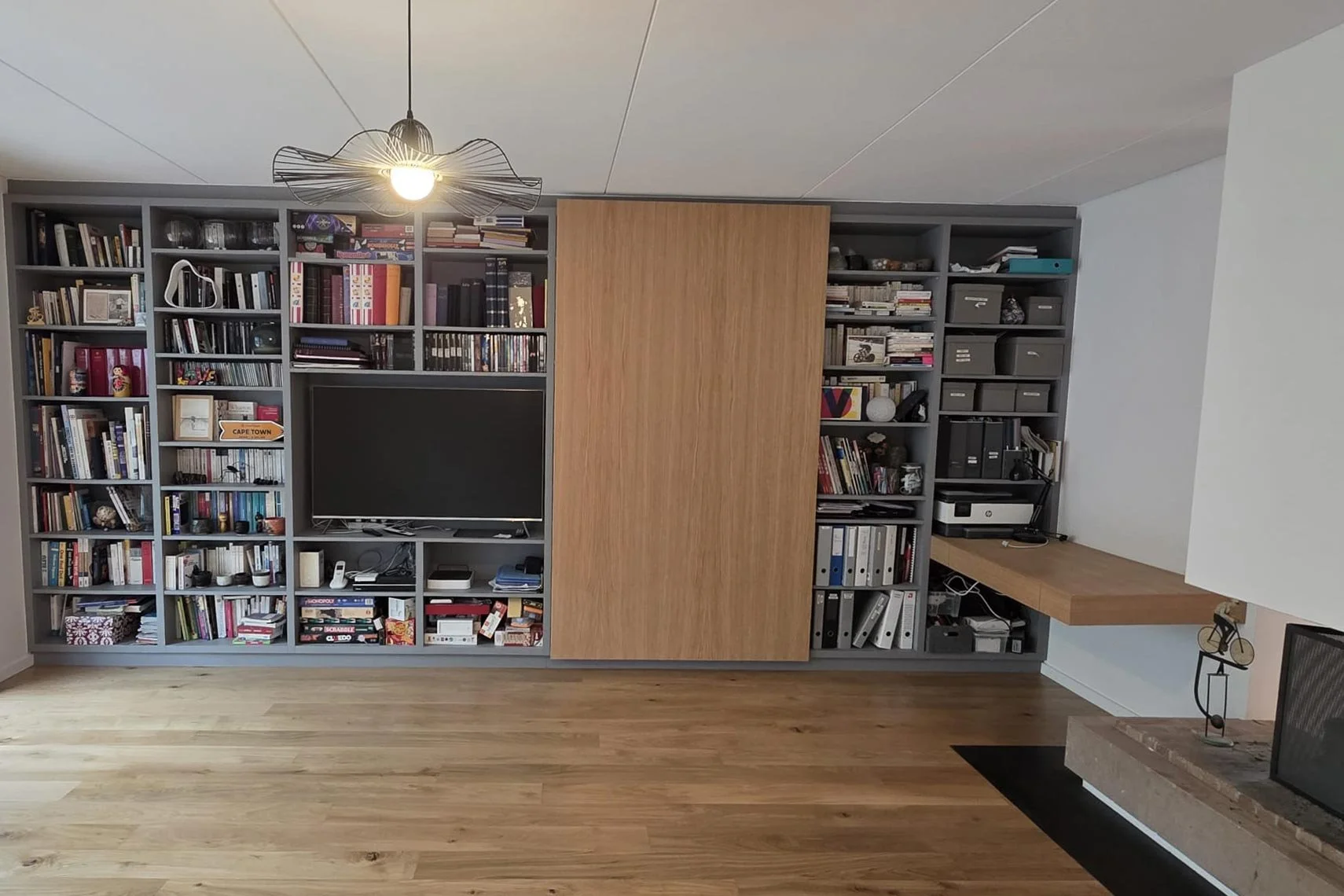 Une bibliothèque murale grise remplie de livres, avec un téléviseur au centre, et un espace de bureau en bois avec une imprimante à proximité. Il y a un plafond avec un luminaire moderne et un parquet en bois naturel.