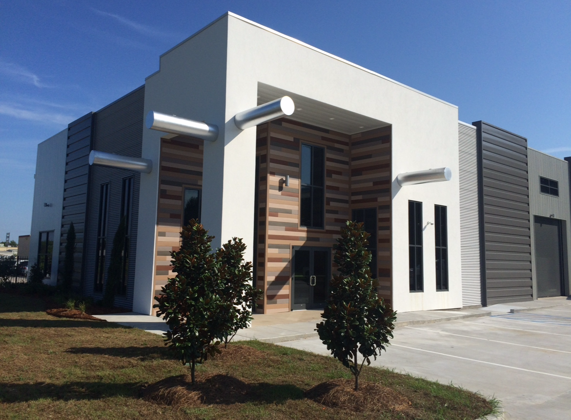 Modern commercial building with white facade, metal pipes extending horizontally, and landscaped area with bushes in front.