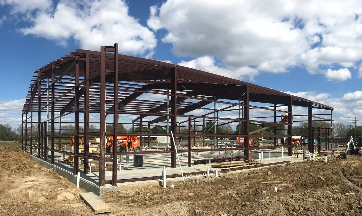 Construction site with a metal building framework being assembled, construction vehicles and equipment visible underneath, and a cloudy sky overhead.