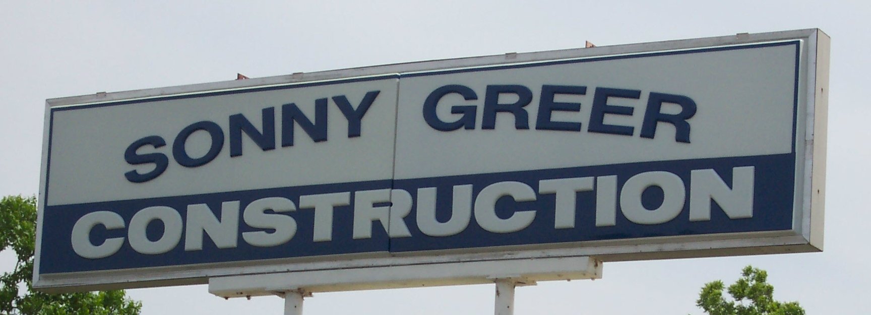A rectangular sign for Sonny Greer Construction with blue text on a white background, mounted on a white pole, with trees and a cloudy sky in the background.