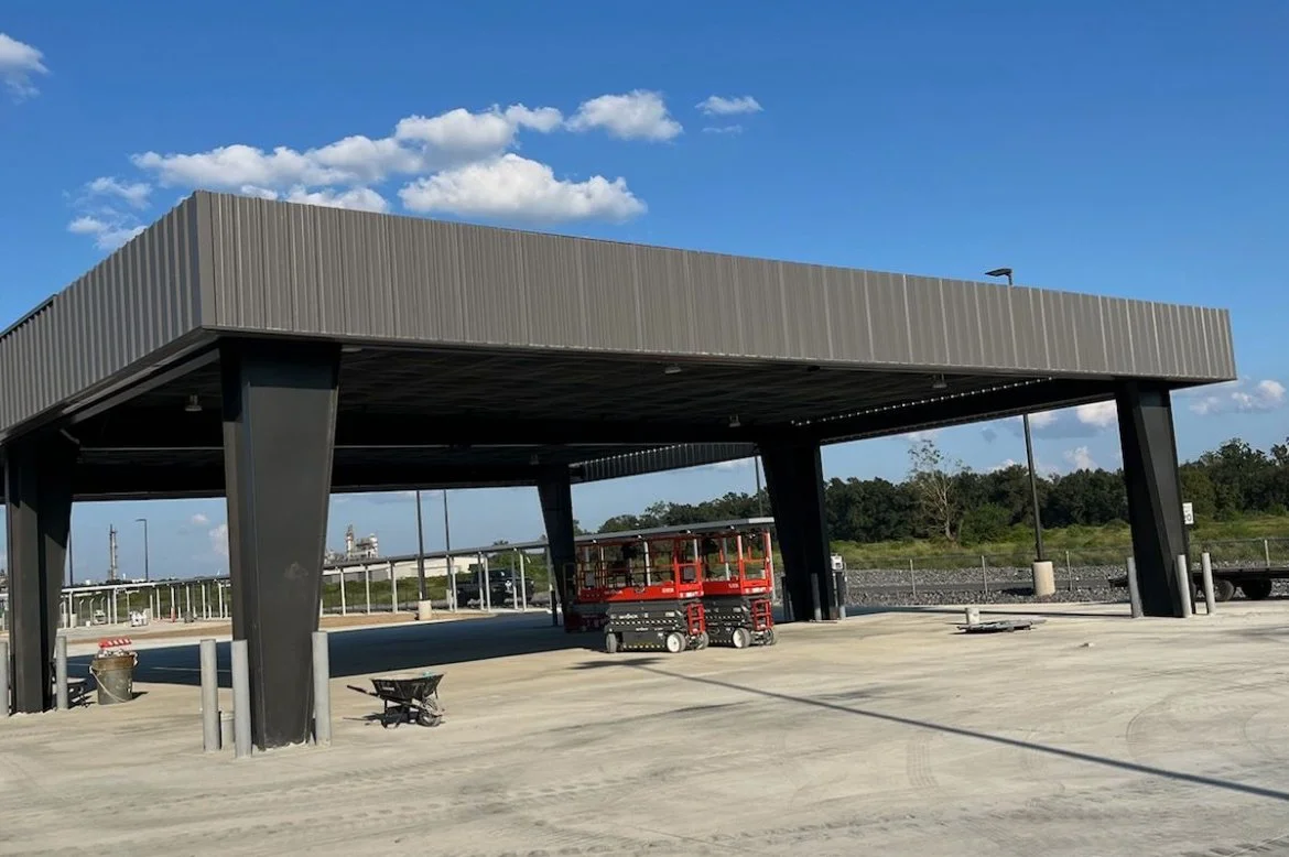 An outdoor construction site with a partially built canopy structure, a red scissor lift, and construction equipment, with a blue sky and some clouds in the background.