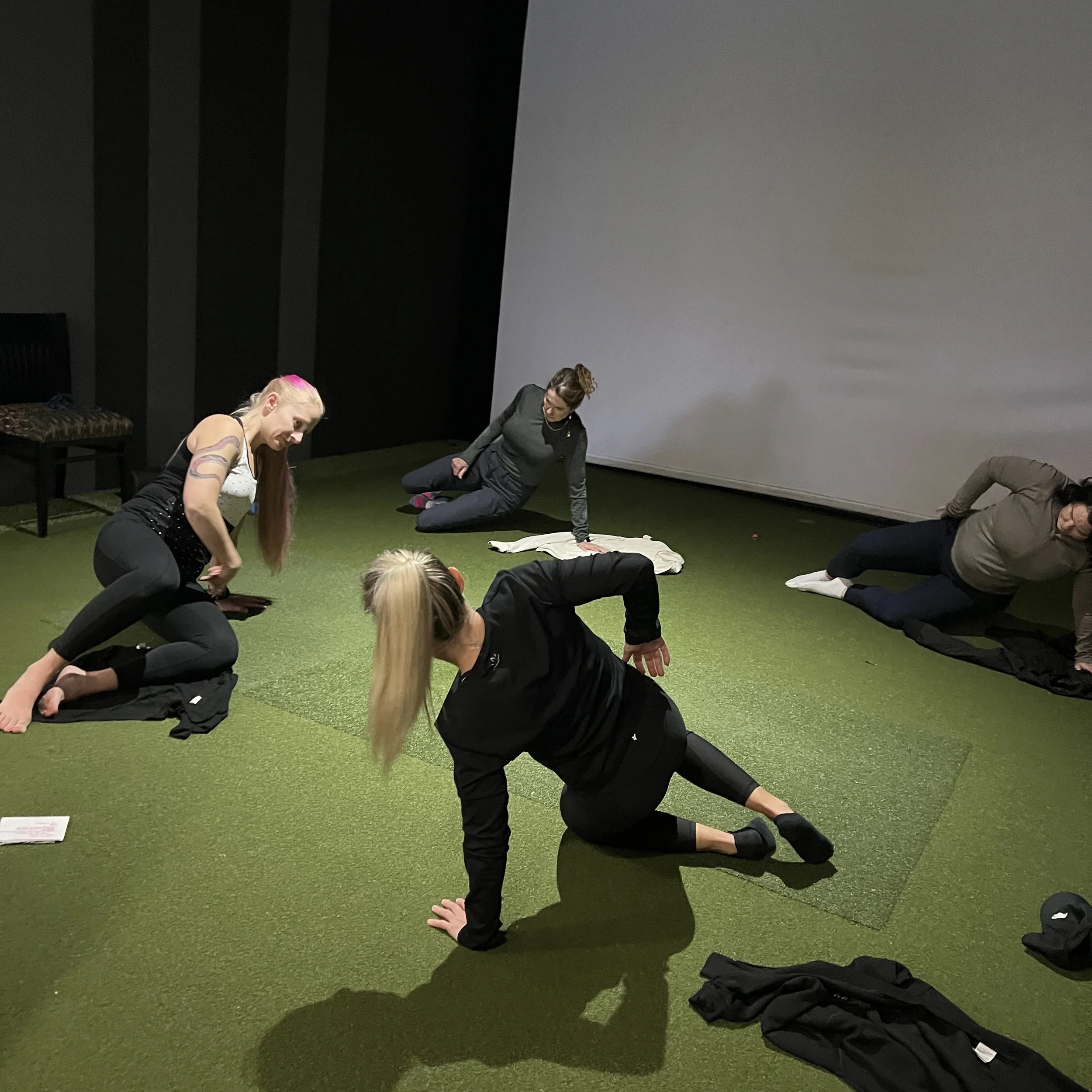 Four women practicing yoga or stretching on a green carpeted floor in a dark room with black walls and a white wall or screen.