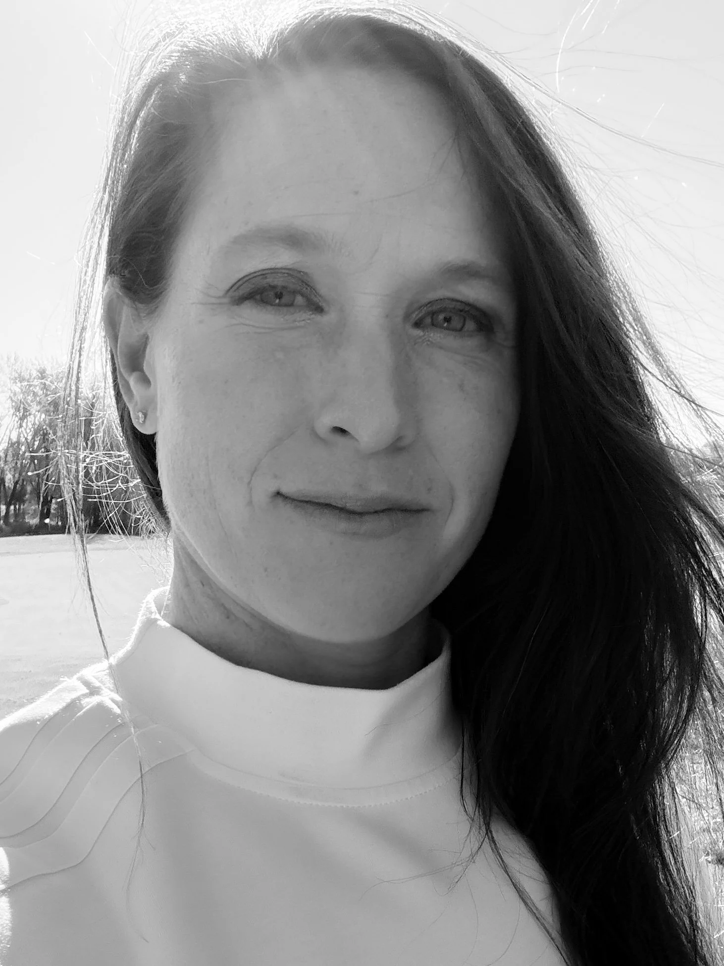 Black and white portrait of a woman with long dark hair, wearing a light-colored shirt, standing outdoors with trees in the background.