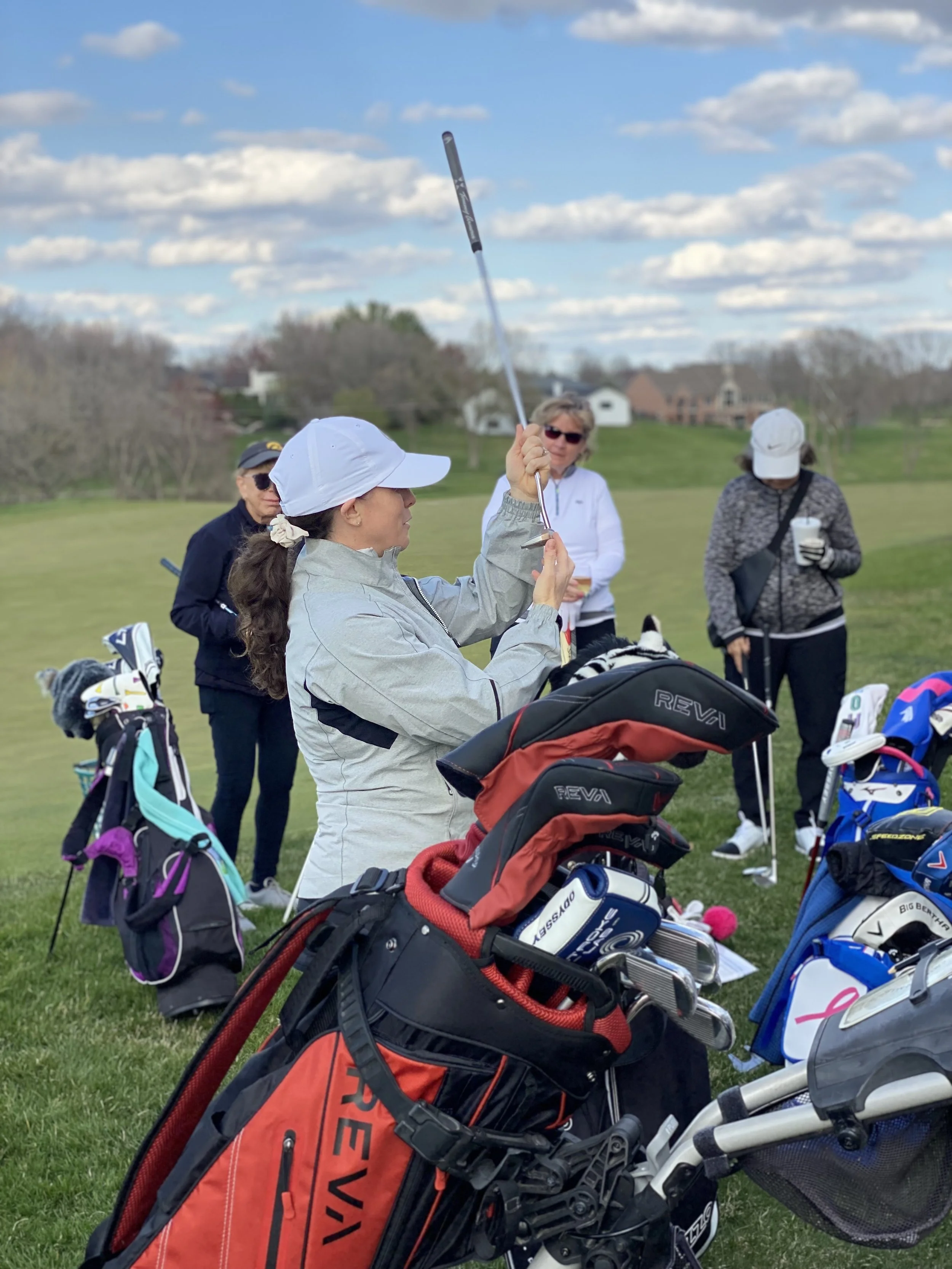 A woman practicing golf on a field with four other women watching, surrounded by golf bags, under a partly cloudy sky.