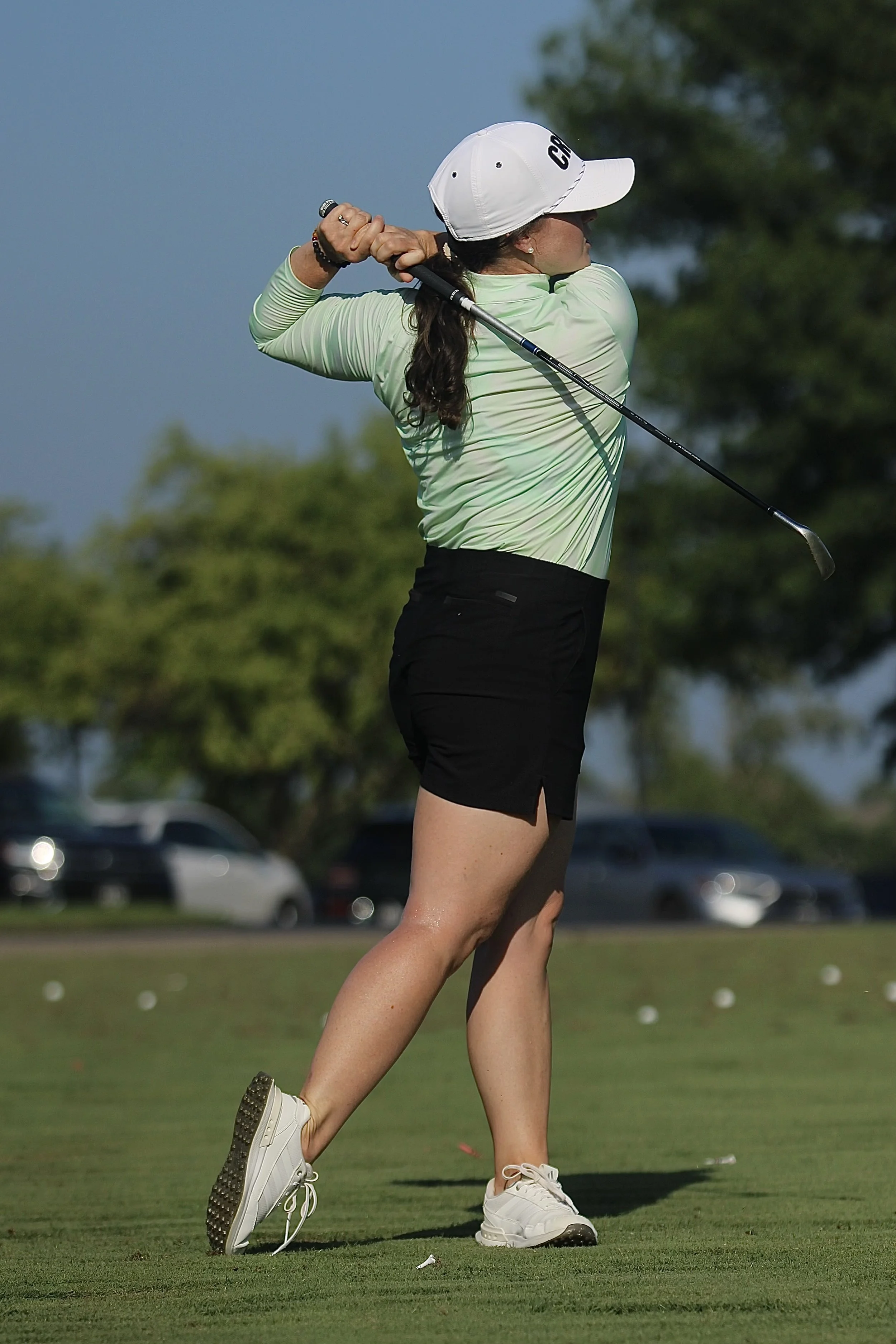 A woman playing golf, swinging a golf club on a golf course with trees and parked cars in the background.