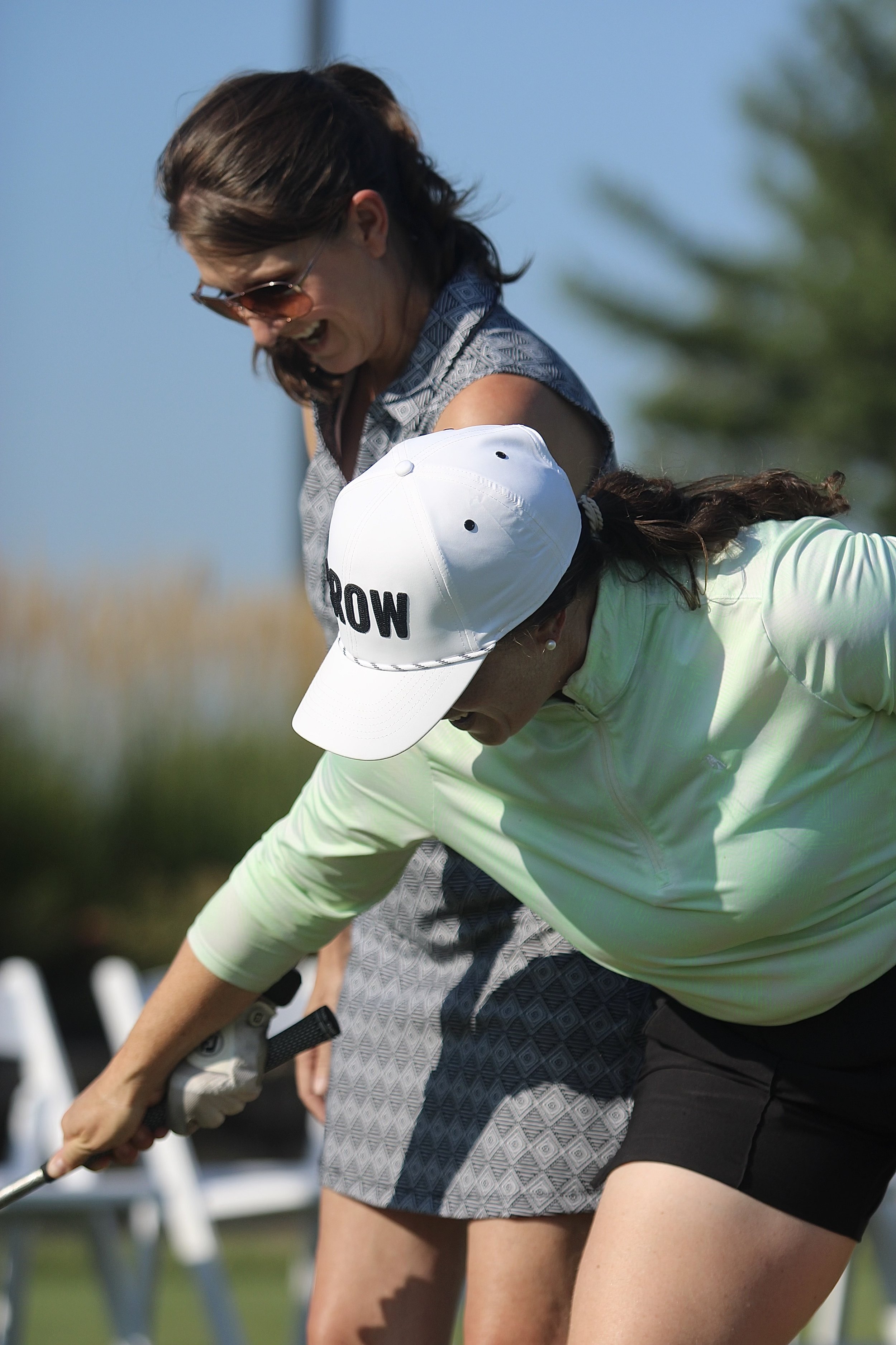 Two women playing golf together, one wearing a white cap and light green shirt, the other with sunglasses and a patterned dress, on a sunny day outdoors.