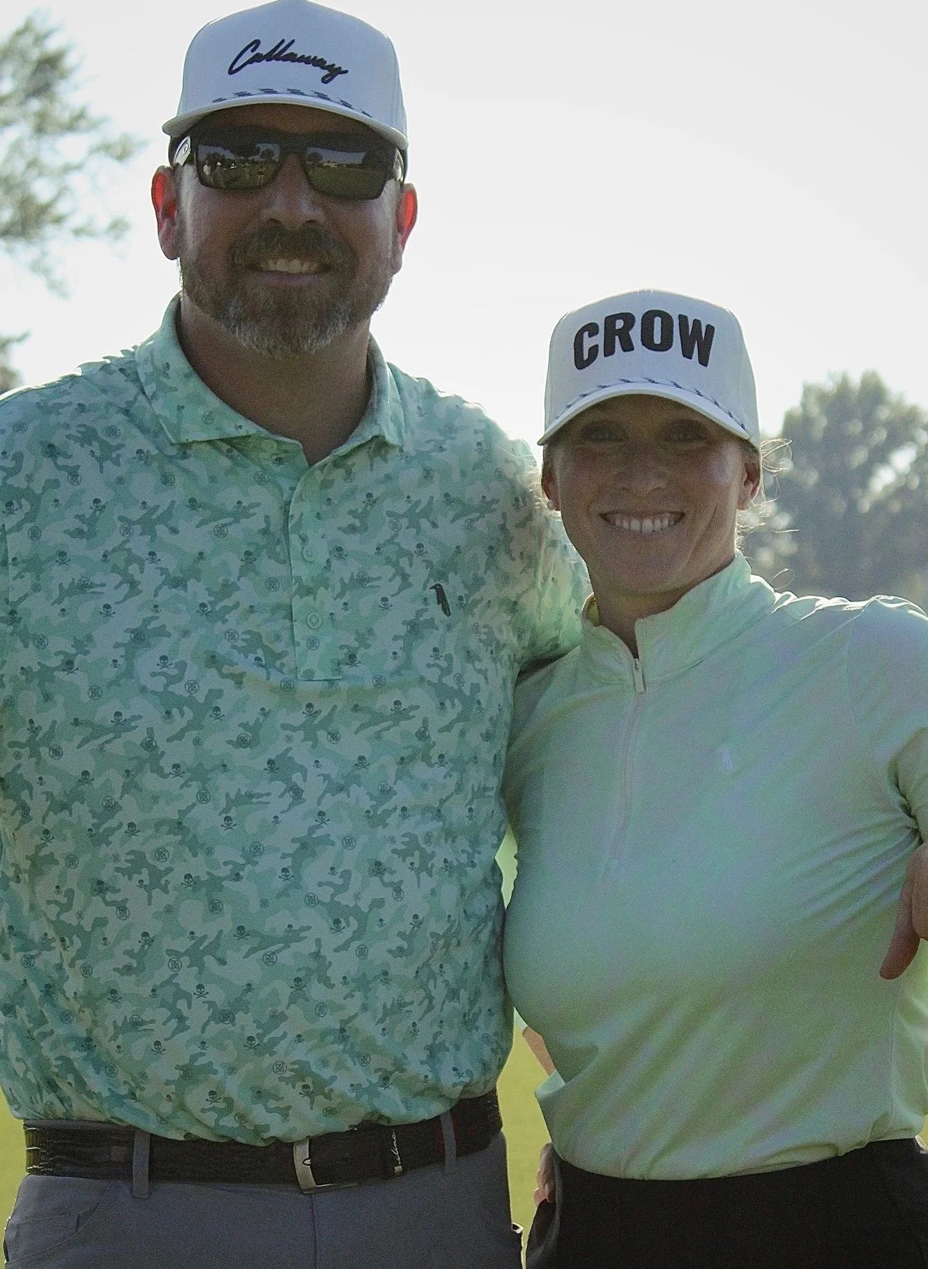 A man and a woman, both smiling, standing outdoors in sunlight. The man has a beard, mustache, sunglasses, a white cap with cursive writing, and a patterned shirt. The woman has a cap with the word 'CROW' and is wearing a light-colored long-sleeve athletic top.