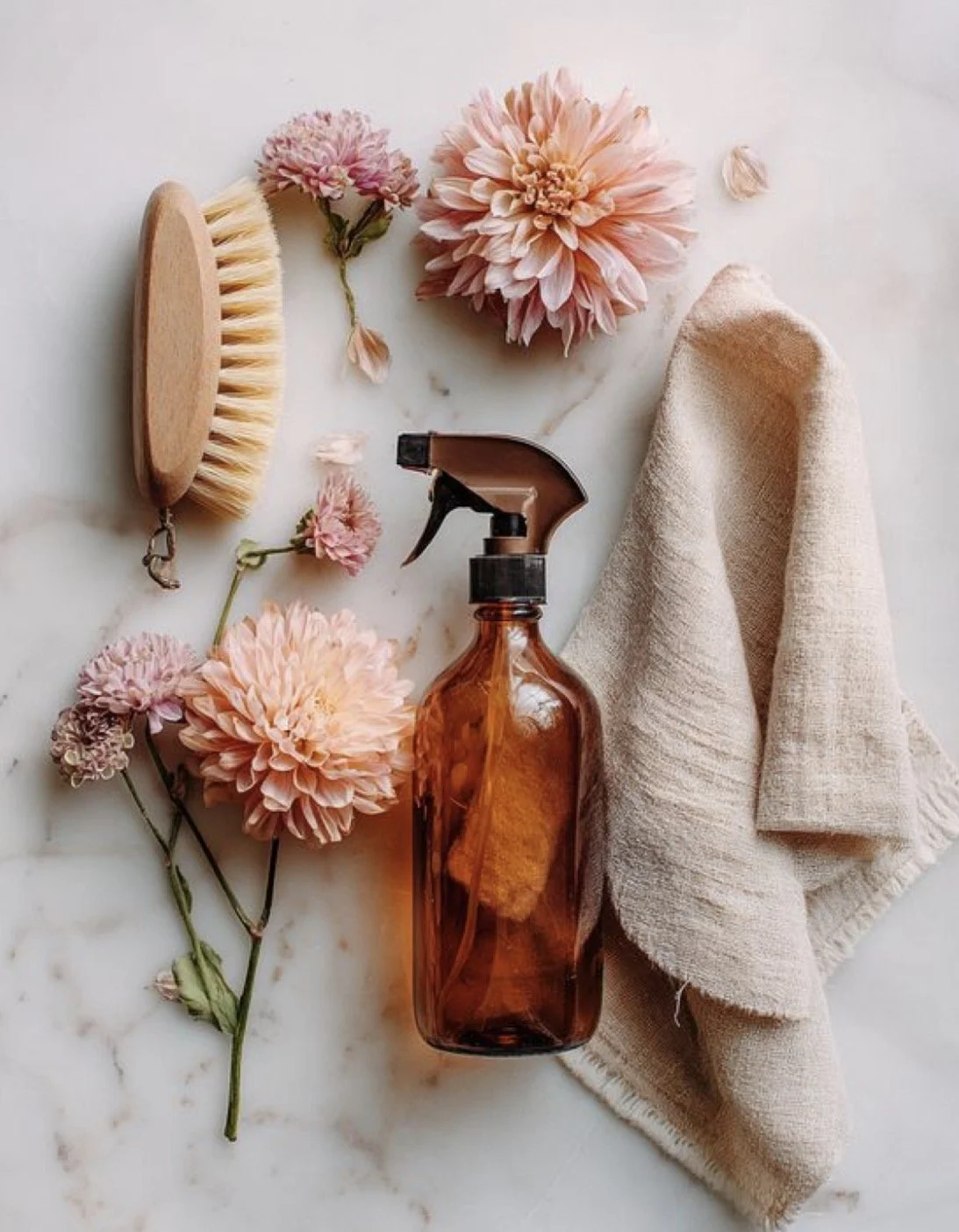A collection of cleaning and grooming items on a marble surface, including a wooden scrub brush, pink flowers, an amber spray bottle with a sponge inside, a beige towel, and scattered flower petals.
