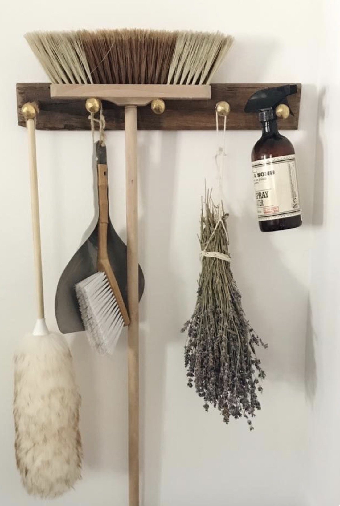 A wall-mounted wooden rack with hooks holding various cleaning and gardening tools, including a broom, dustpan, sprayer, and a bunch of dried lavender.