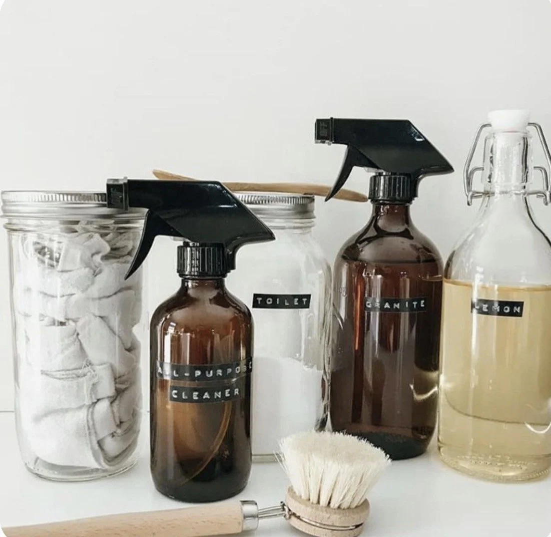 Cleaning supplies including spray bottles labeled 'ALL PURPOSE CLEANER' and 'TOILET', a jar of cloths, a jar of baking soda, a bottle of glass cleaner, a lemon-scented spray, a scrub brush, and a wooden scrubber on a white surface.