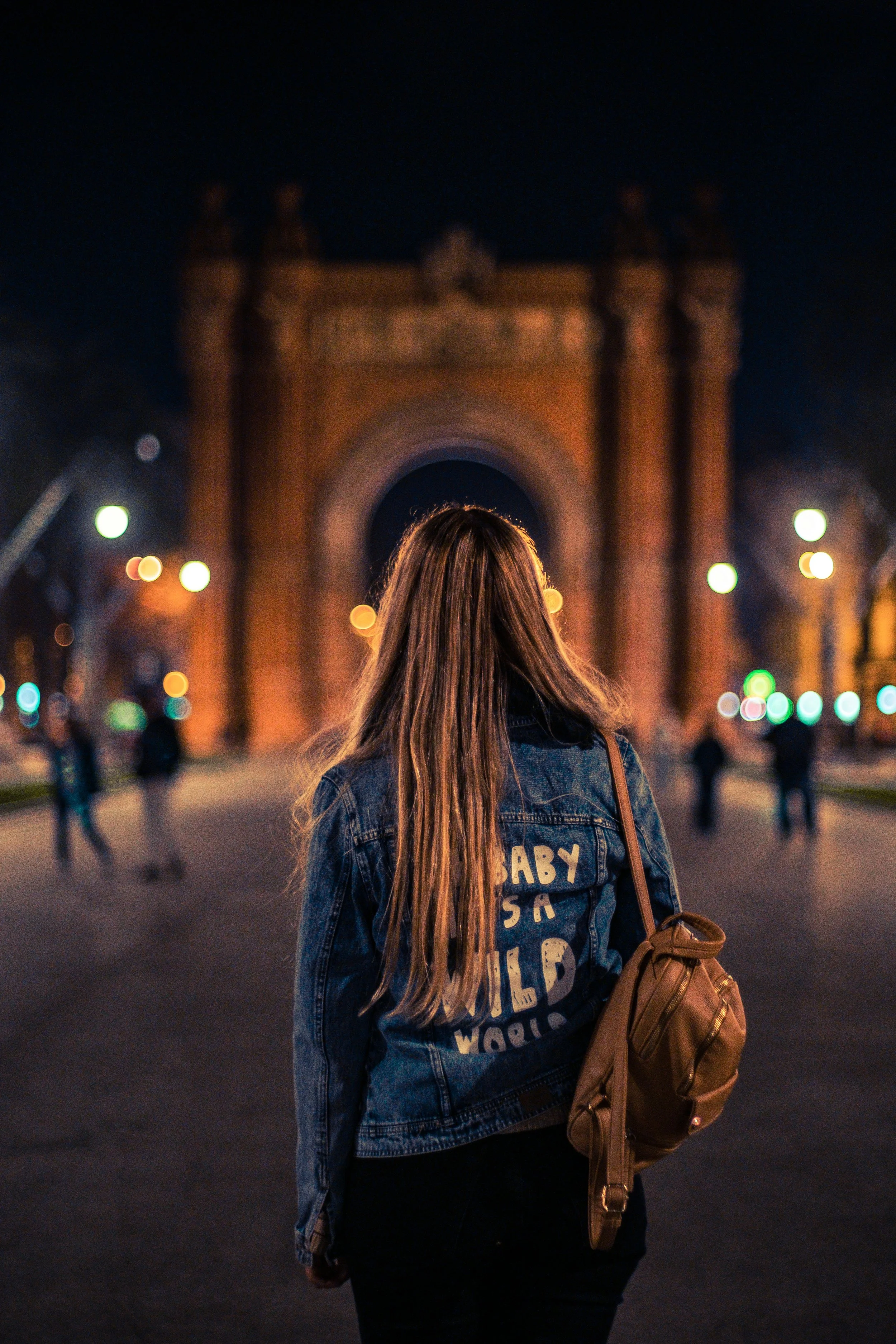 Joven con chaqueta de mezclilla y mochila observando el Arco de Triunfo en París, iluminado por la noche.