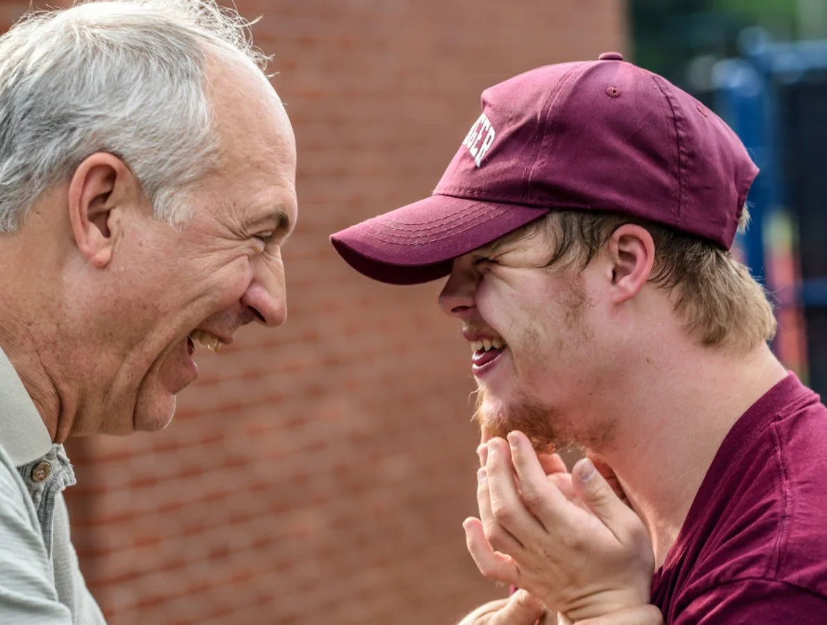 An older man and a young man are smiling and laughing face to face outdoors, with a brick wall in the background.