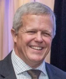 A smiling man with gray hair wearing a suit and tie at an indoor event.