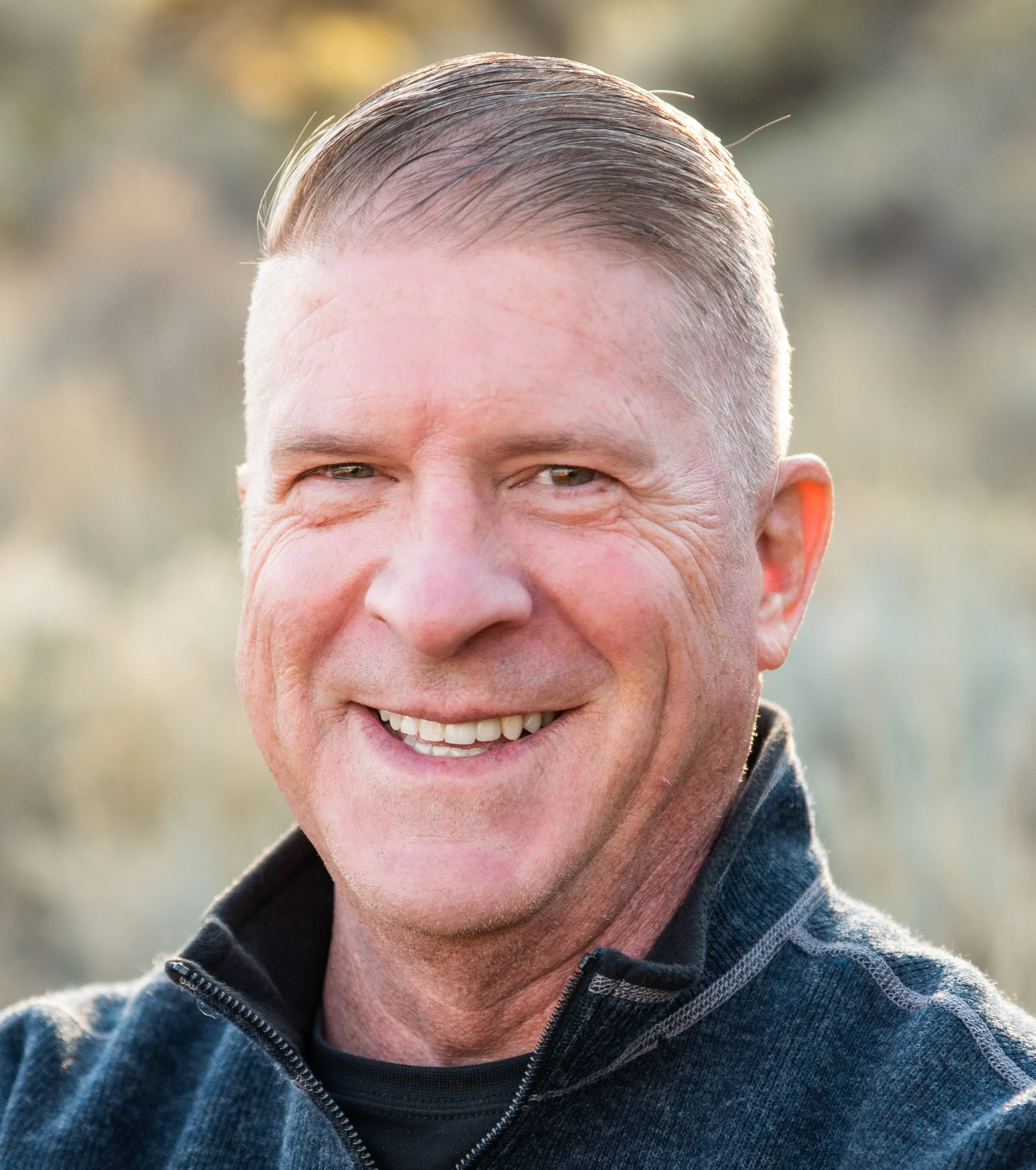 A close-up portrait of a smiling middle-aged man with short, combed-back hair, wearing a dark zip-up jacket outdoors in natural light.