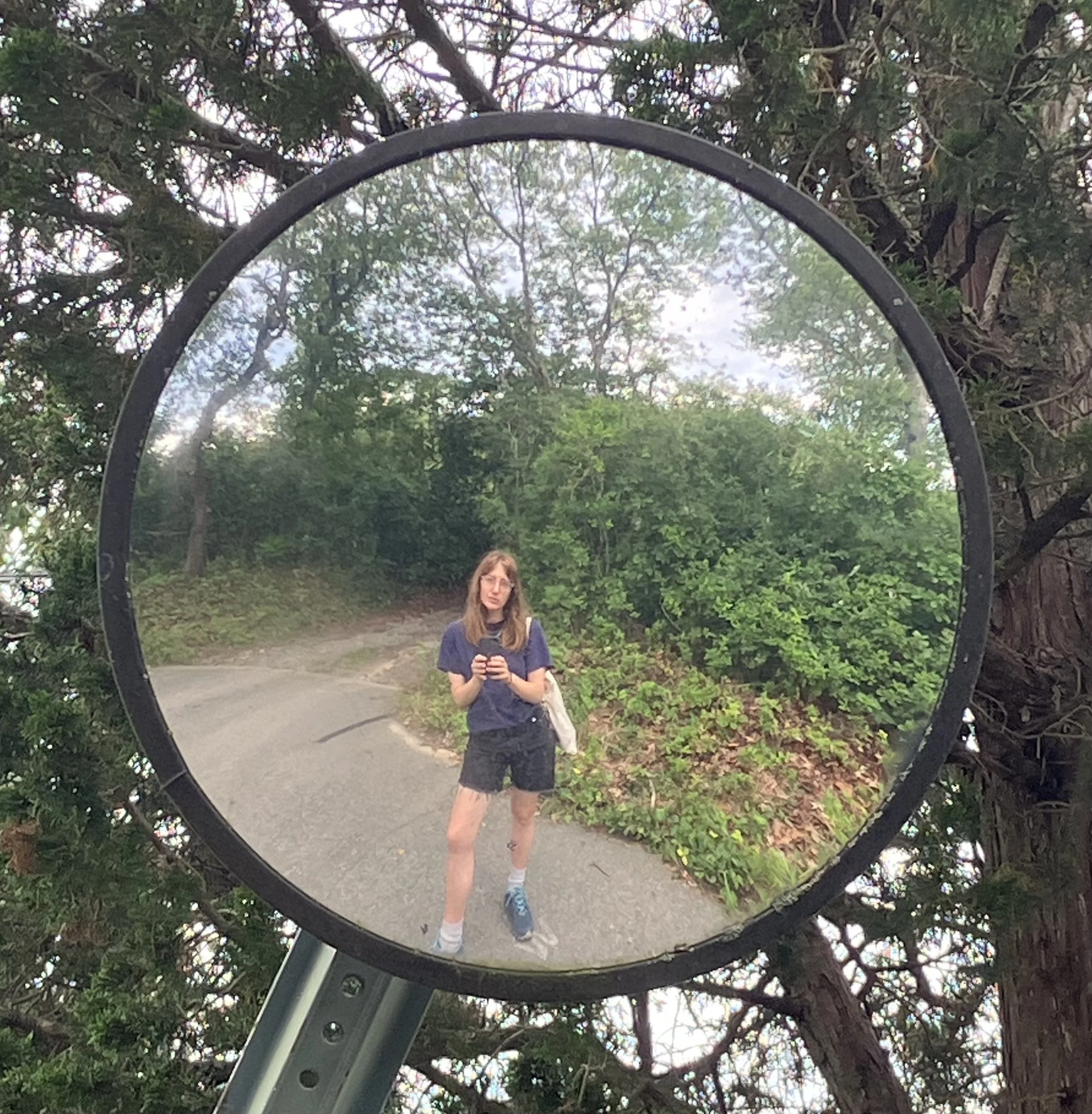A woman taking a selfie with a smartphone in a circular mirror outdoors, surrounded by green trees and bushes, on a cloudy day.