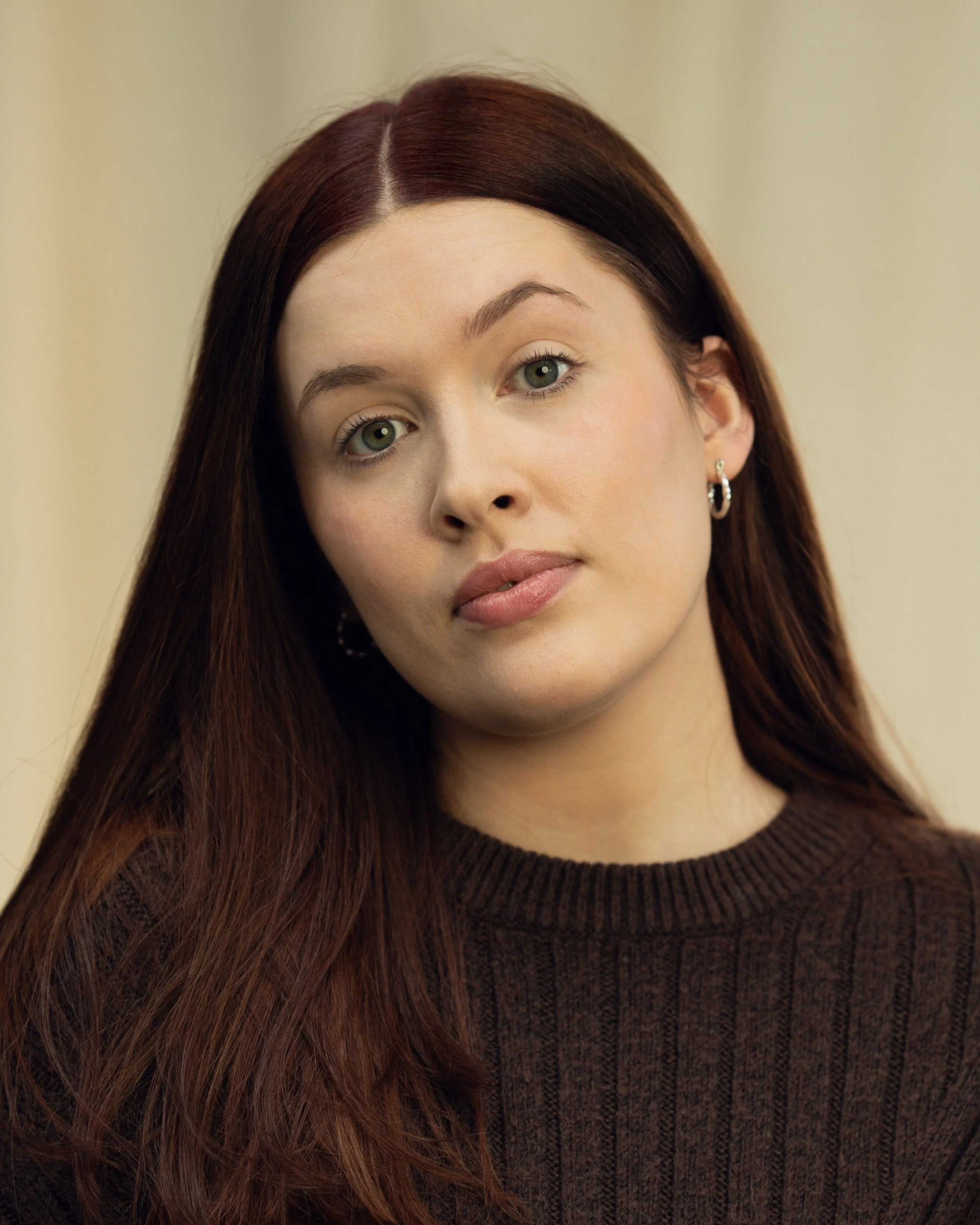 A woman with long red hair and green eyes, wearing a dark brown sweater and earrings, looking at the camera.
