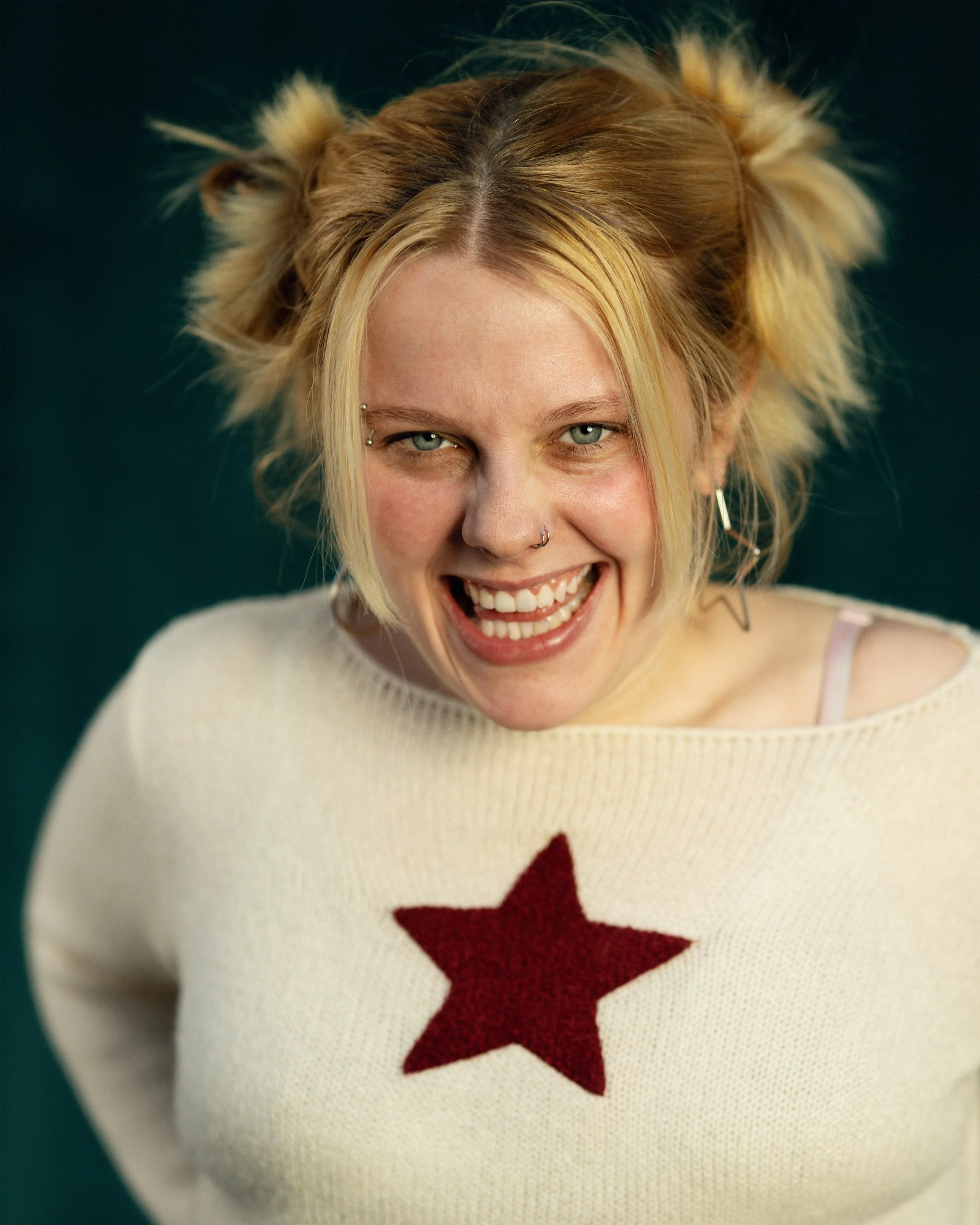 A young woman with blonde hair styled in pigtails, smiling and showing teeth, wearing a cream sweater with a red star on the front, earrings, and facial piercings against a dark background.