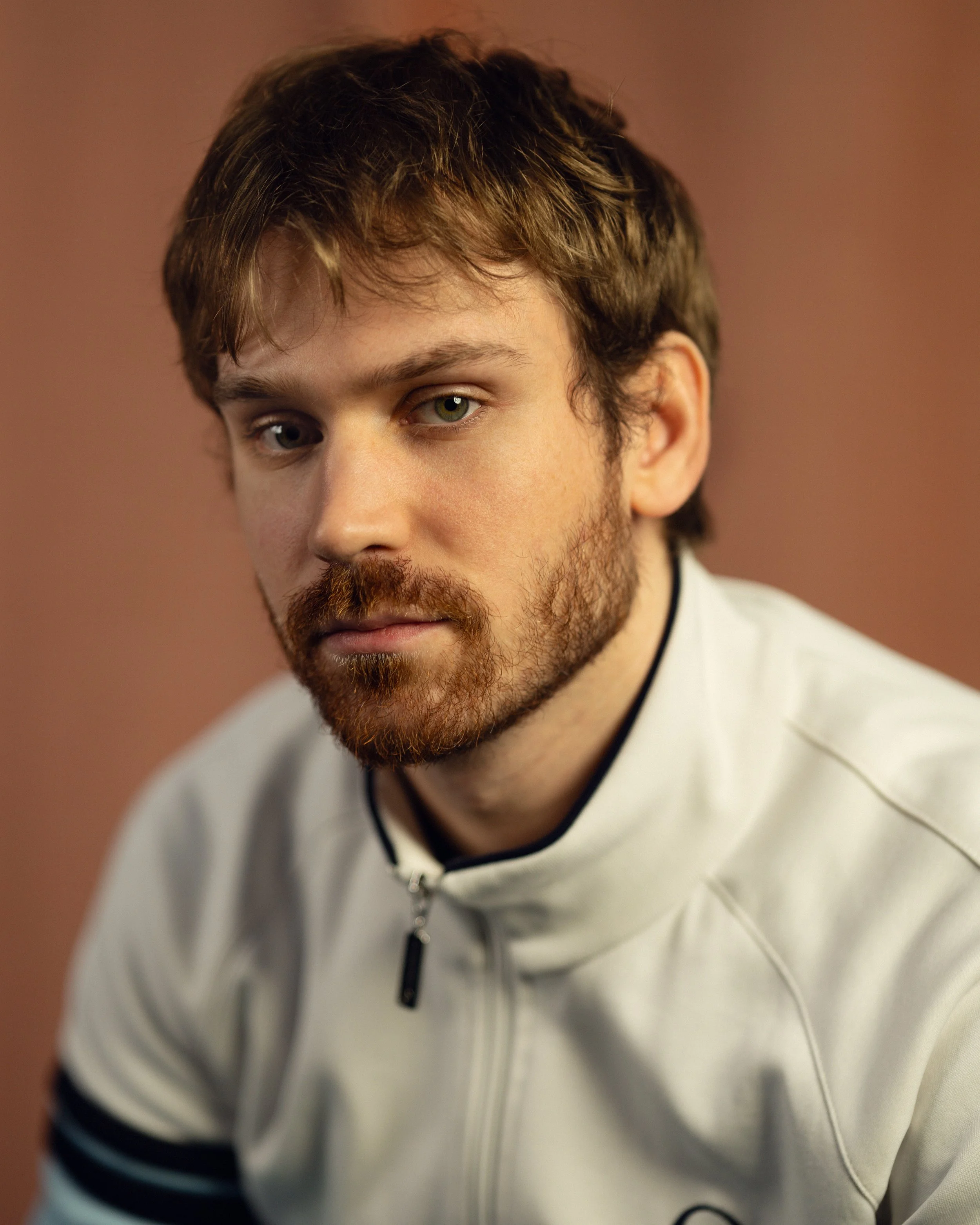 A young man with light brown hair and a reddish beard, wearing a white sports jacket, looking slightly to the right with a serious expression against a plain brown background.