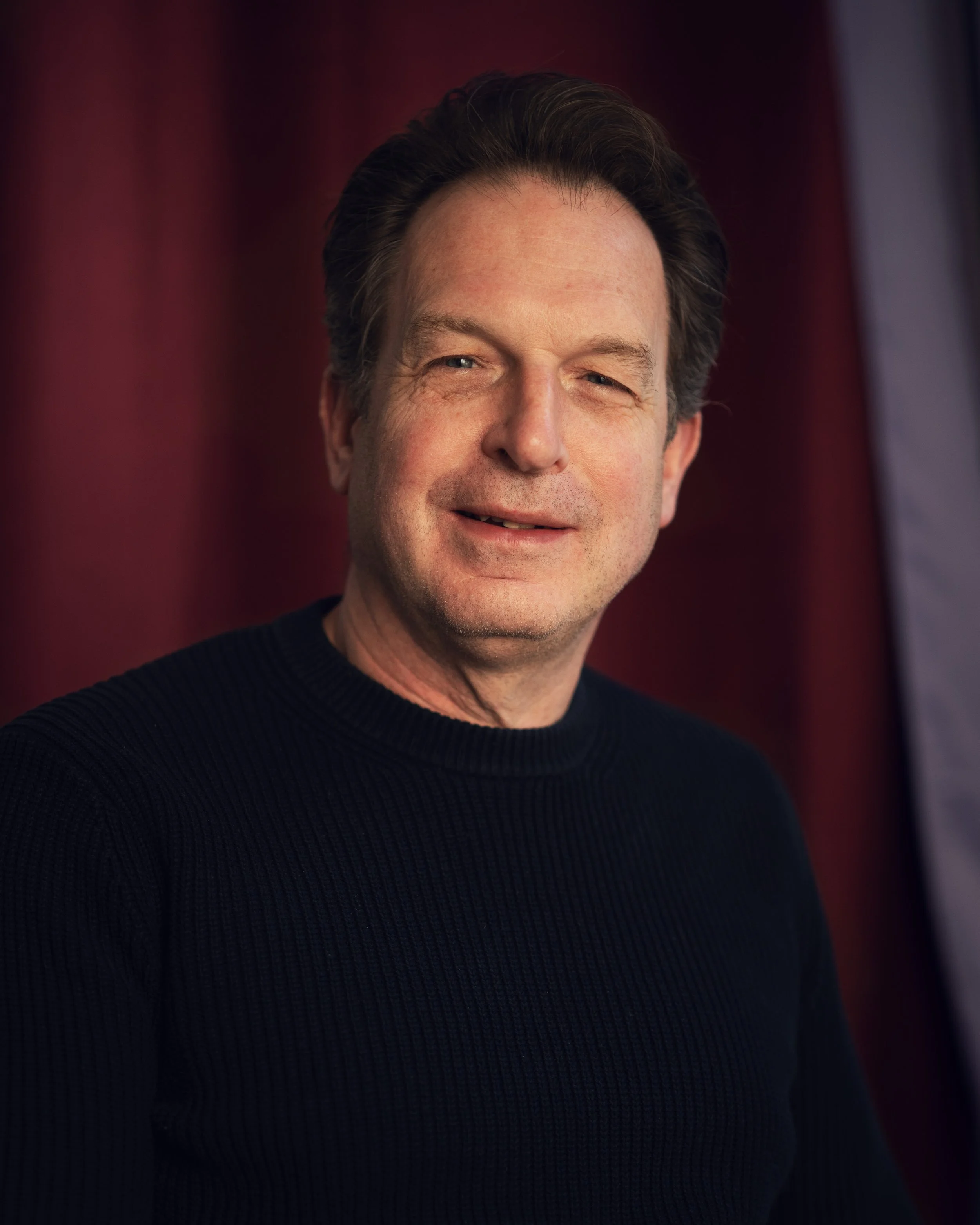 A headshot of man with dark hair and a fair complexion, smiling, wearing a black ribbed sweater, in front of a blurred background with red and dark shades.