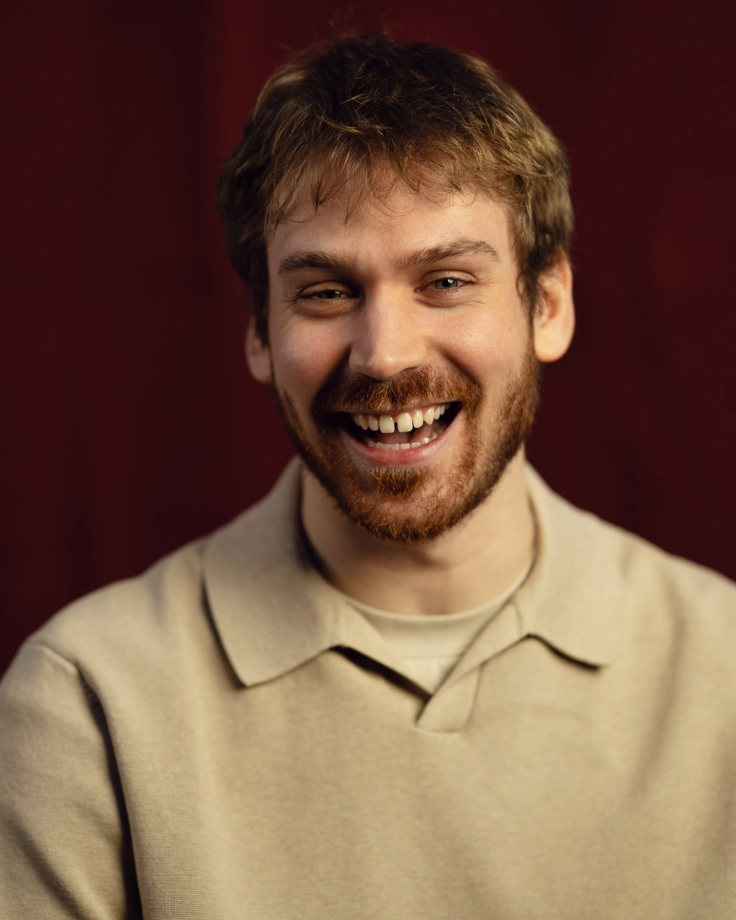 A smiling man with light skin, brown hair, and a beard, wearing a beige collared shirt, against a dark red background.