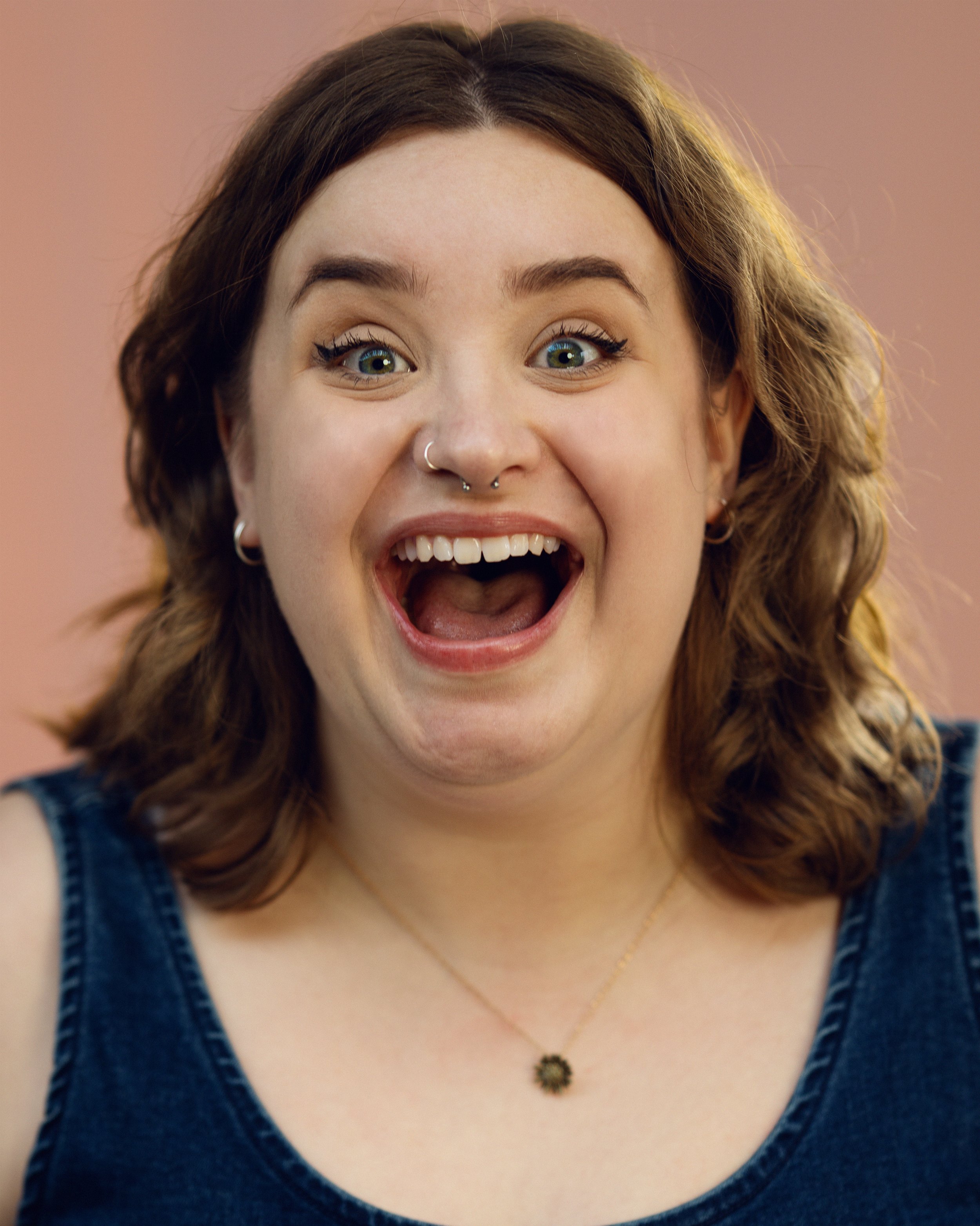 An actress with shoulder-length brown hair, green eyes, smiling widely with an open mouth, wearing a blue tank top and a gold necklace, against a pink background.