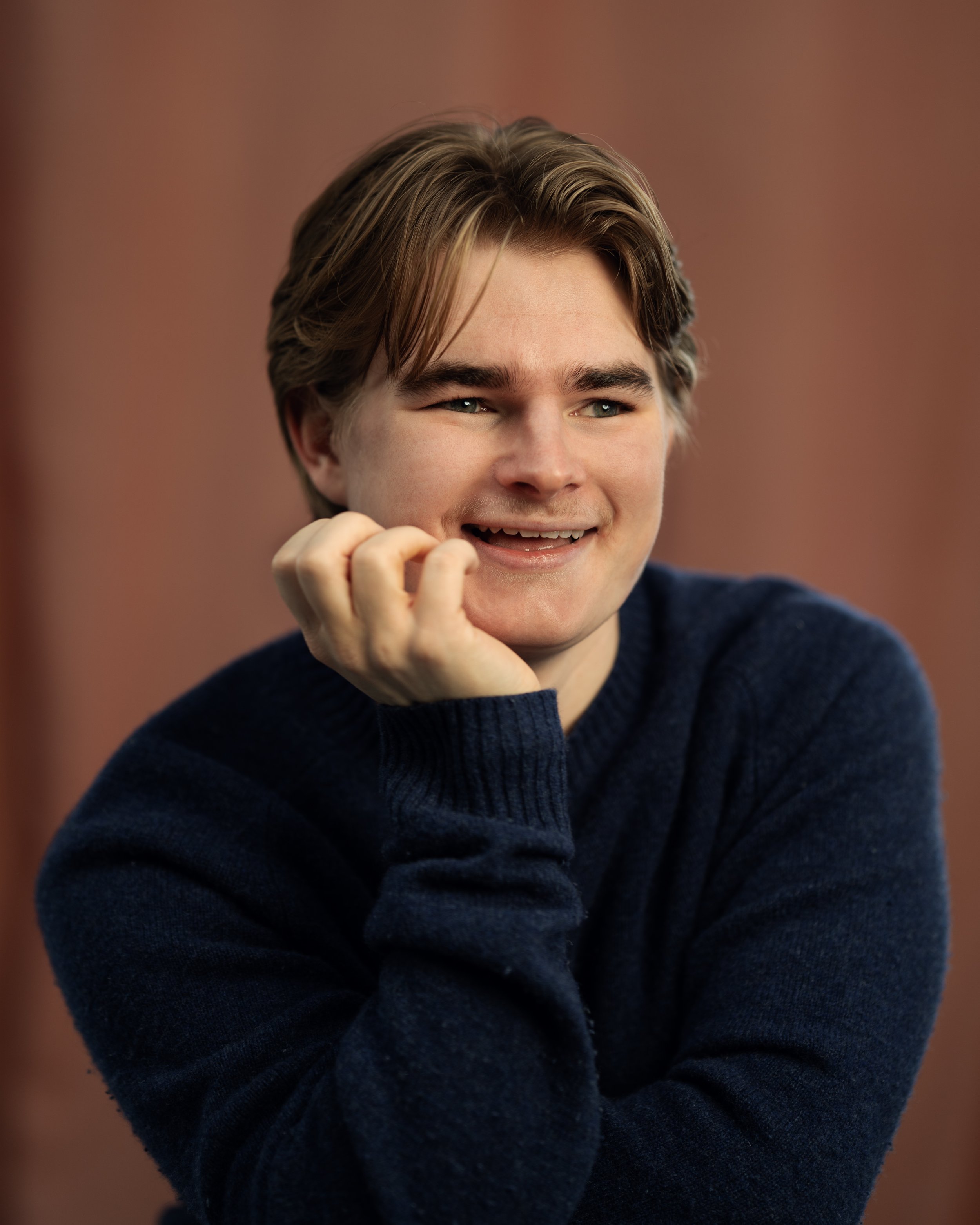 A young man with light brown hair and blue eyes, wearing a dark blue sweater, smiling with his hand resting on his cheek, against a blurred brownish background.