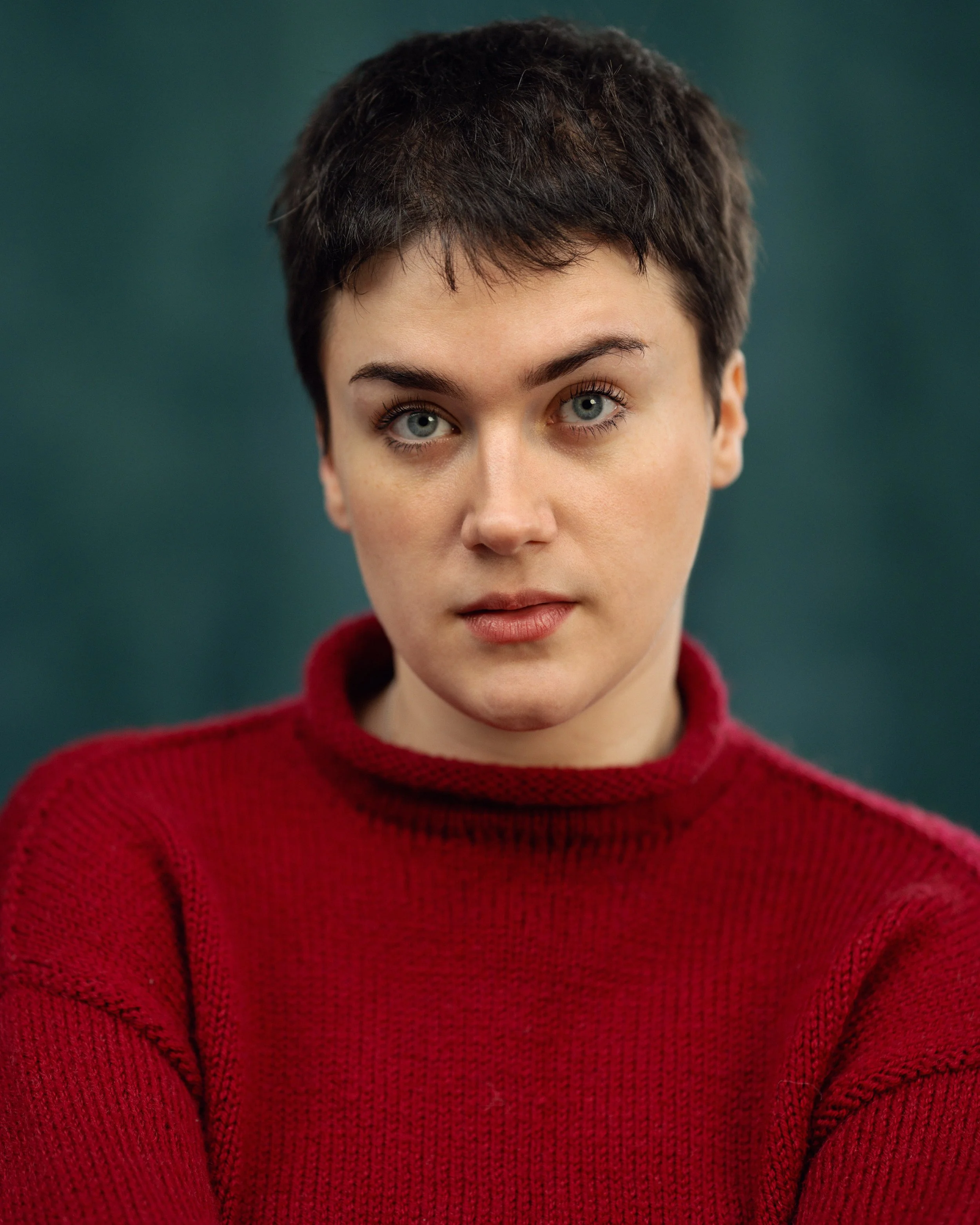 A woman with short brown hair and blue eyes wearing a red sweater against a dark green background.