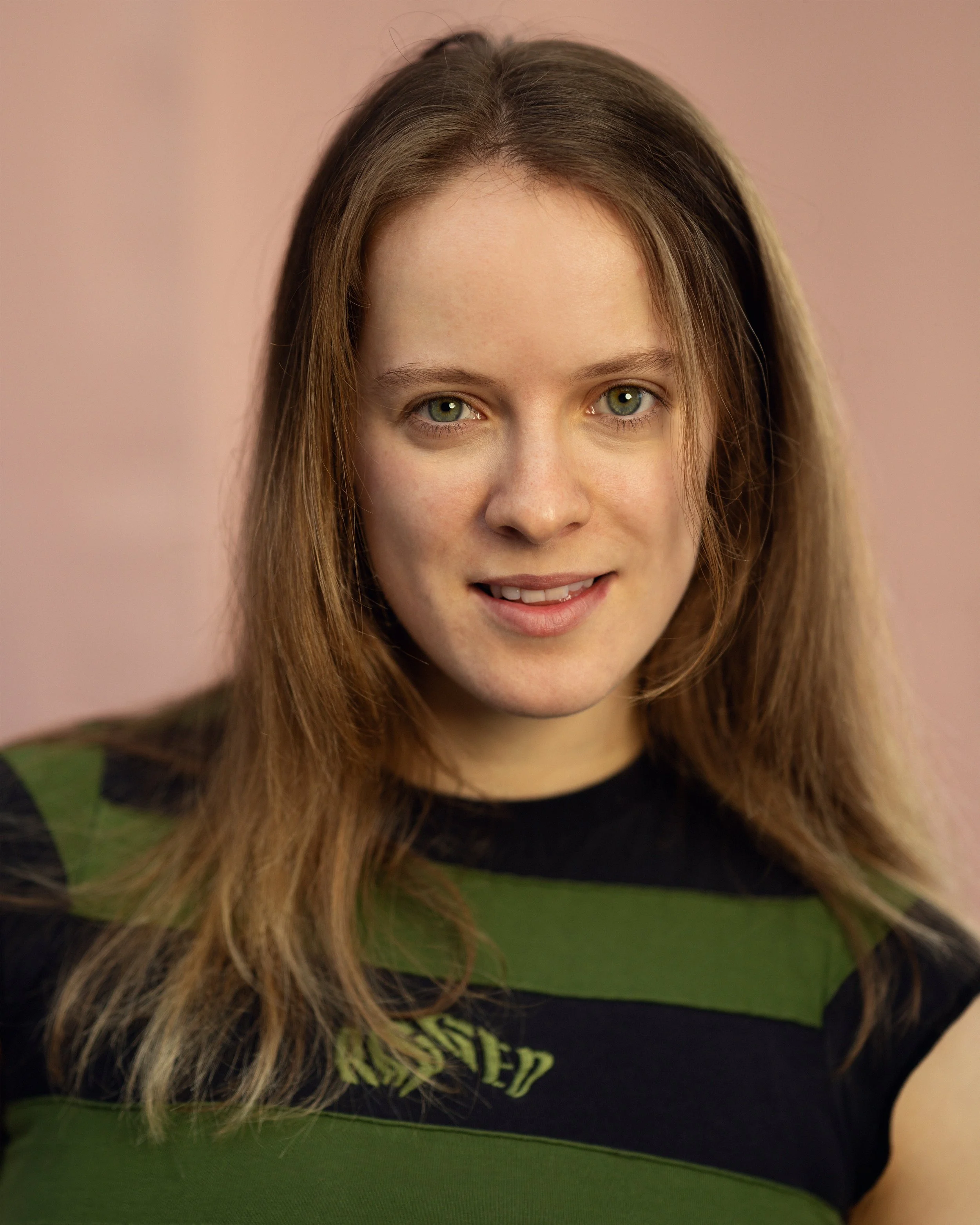 A woman with long brown hair, light skin, and green eyes smiling slightly, wearing a black and green striped shirt, against a pinkish background.