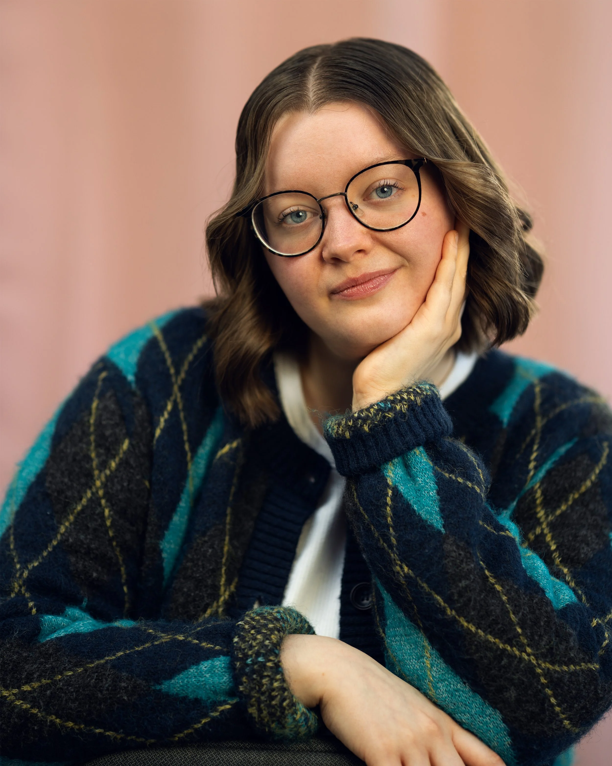 A young woman with shoulder-length brown hair wearing glasses, a white shirt, and a dark plaid sweater, resting her face on her hand and smiling softly, against a pink background.