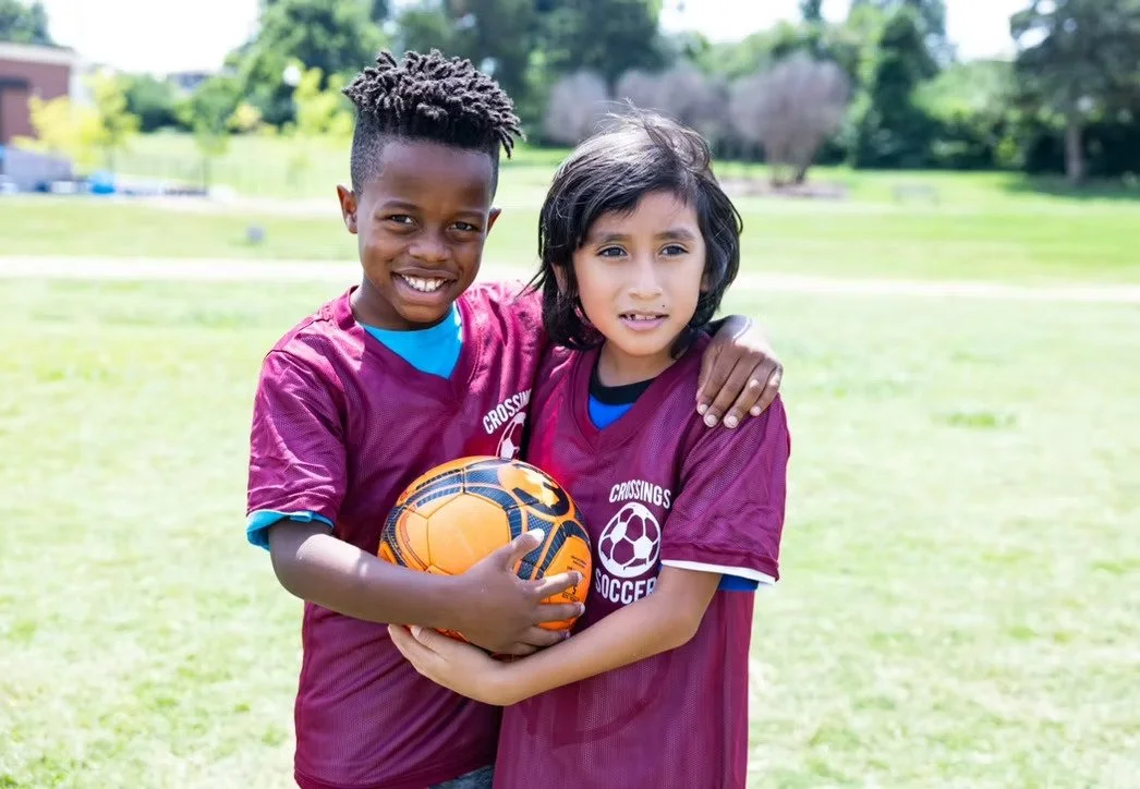 Two children wearing maroon Crossings Soccer jerseys standing outdoors on a soccer field. They are holding a yellow and black soccer ball, smiling, with one child's arm around the other's shoulder.
