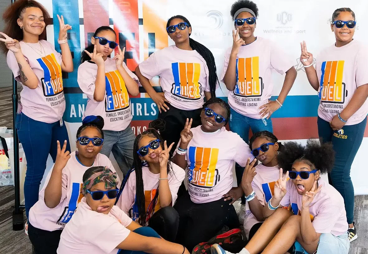 Group of young girls posing for a photo, wearing matching pink T-shirts with a colorful logo and sunglasses, in front of a banner that reads 'Girls' and 'Education'.