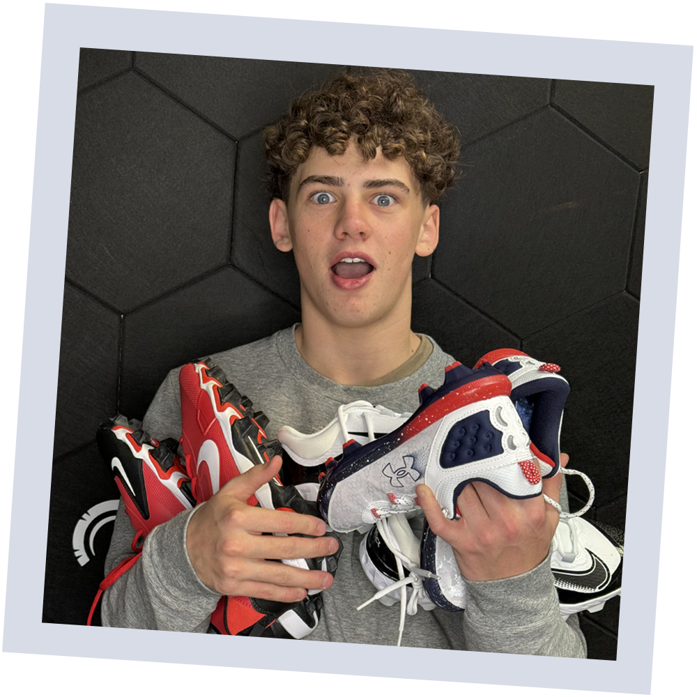 Young man with curly hair holding multiple athletic shoes, looking surprised or excited, sitting against a black padded wall.