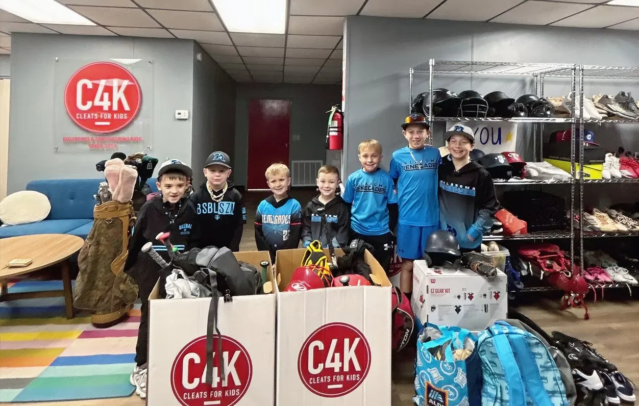 Group of young boys in hockey gear standing inside a sport facility with shelves of helmets, shoes, and sports equipment behind them, and large boxes with cleat for kids branding in the foreground.