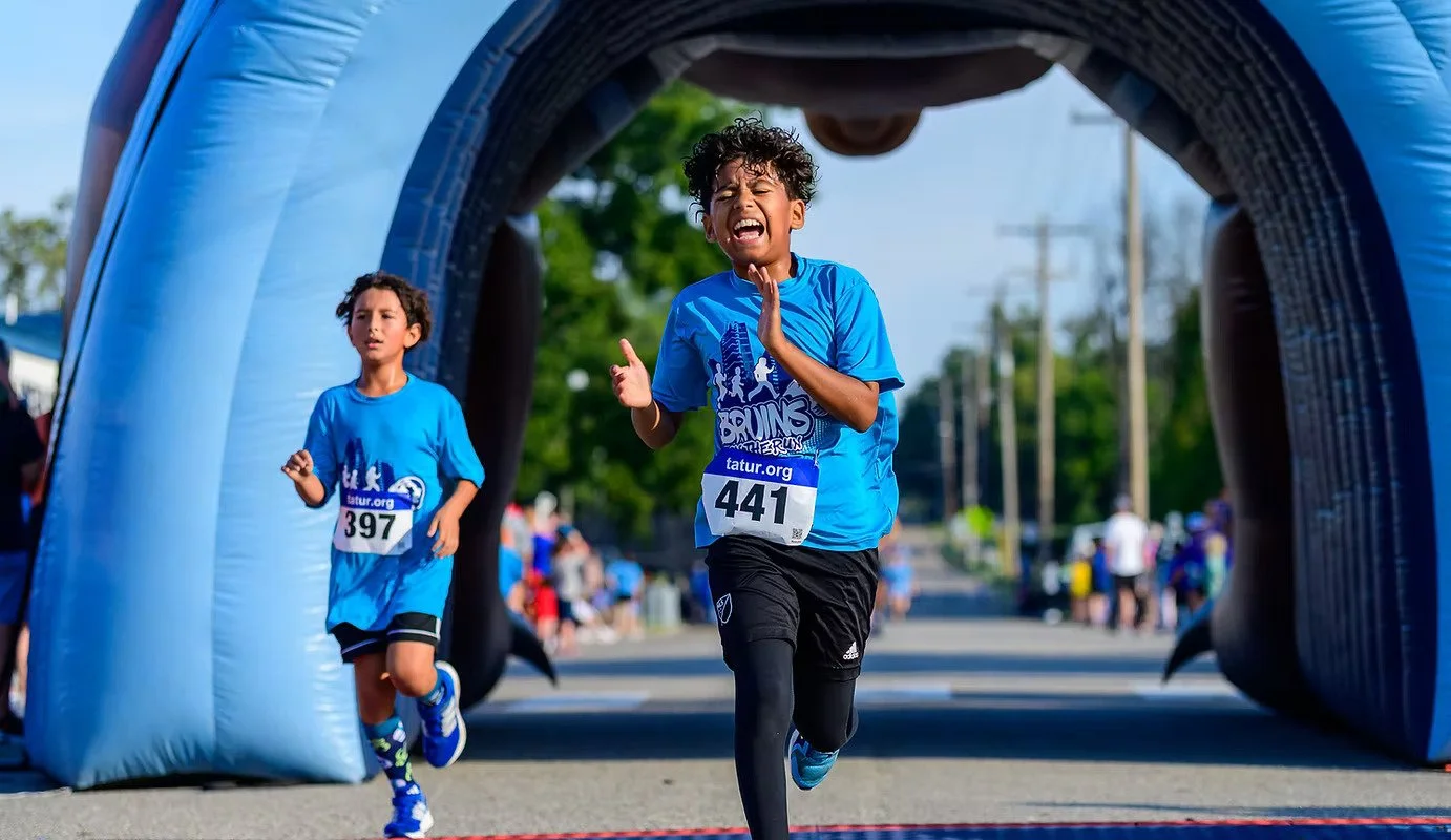 Two children race through an inflatable arch during a fun run, wearing blue shirts, with one child visibly emotional and the other child focused.