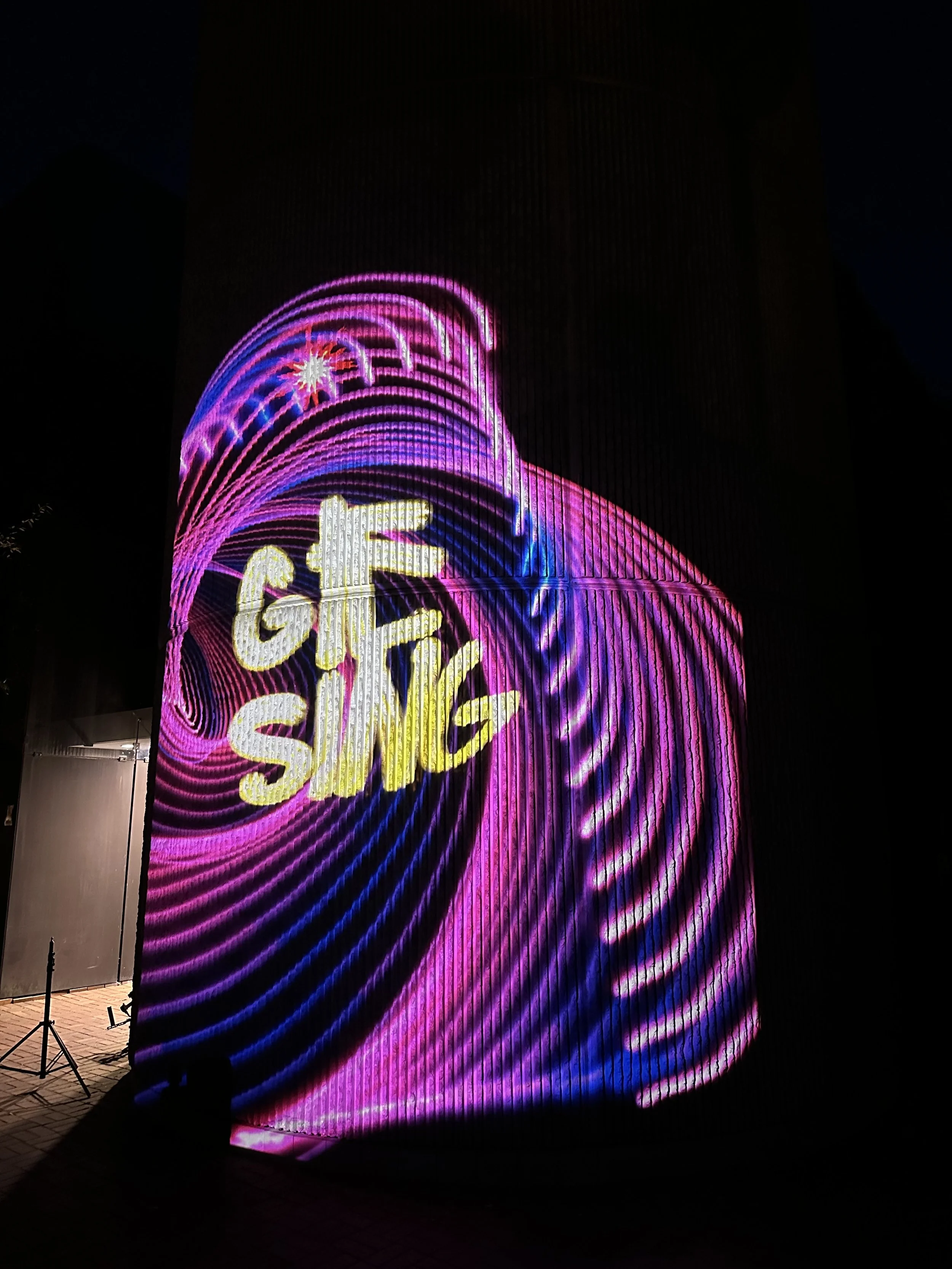 Leuchtreklame mit den Worten "GIRL SING" auf einer dunklen Gebäudefassade bei Nacht, in Neonfarben pink, blau und gelb, mit spiralförmigen Lichtmustern.