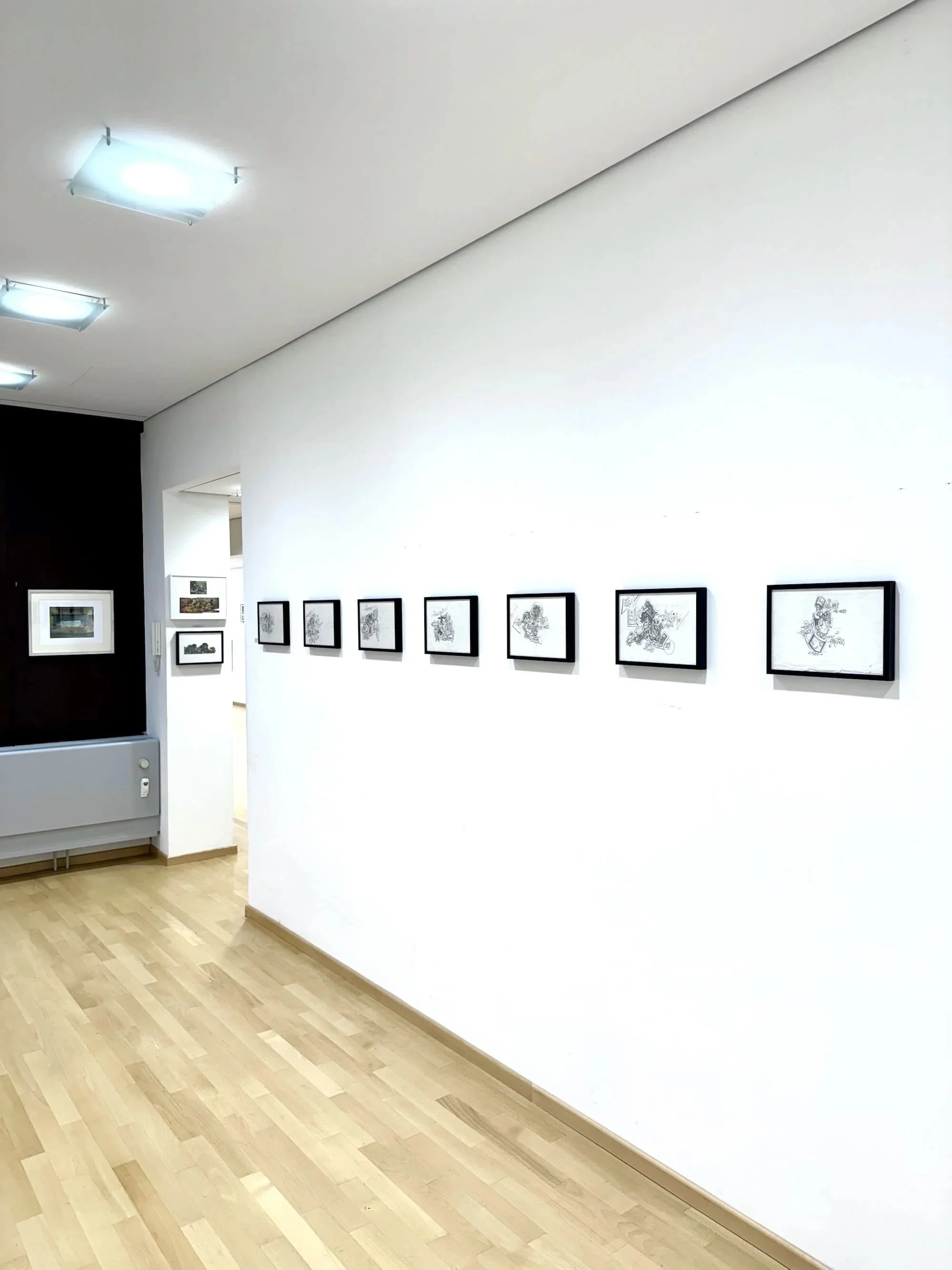 Eine Kunstgalerie mit weißen Wänden, schwarz gerahmten Zeichnungen entlang der Wand, hellem Holzboden, Deckenleuchte und einem Übergang zu einem anderen Raum mit weiteren Kunstwerken.
