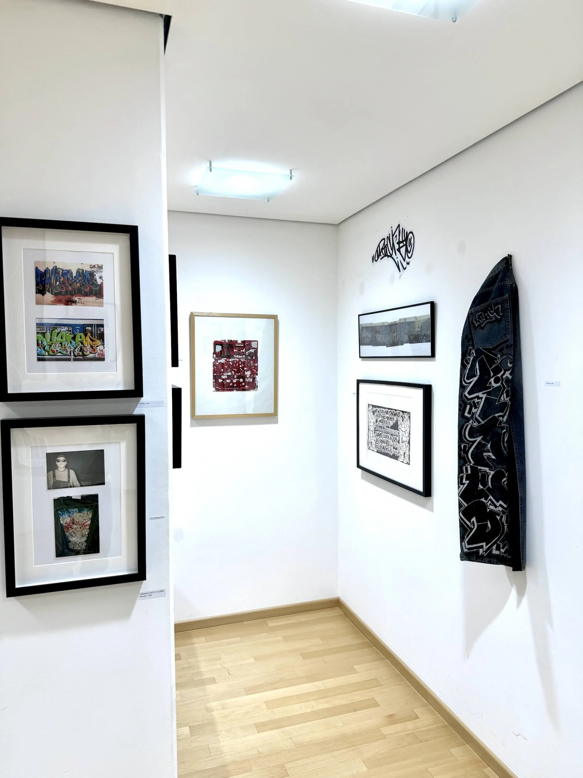 Eine Kunstgalerie mit Bildern an weißen Wänden, darunter Graffiti-Kunst, Schwarz-Weiß-Fotografien und ein schwarzes Tuch mit Graffiti-Design.