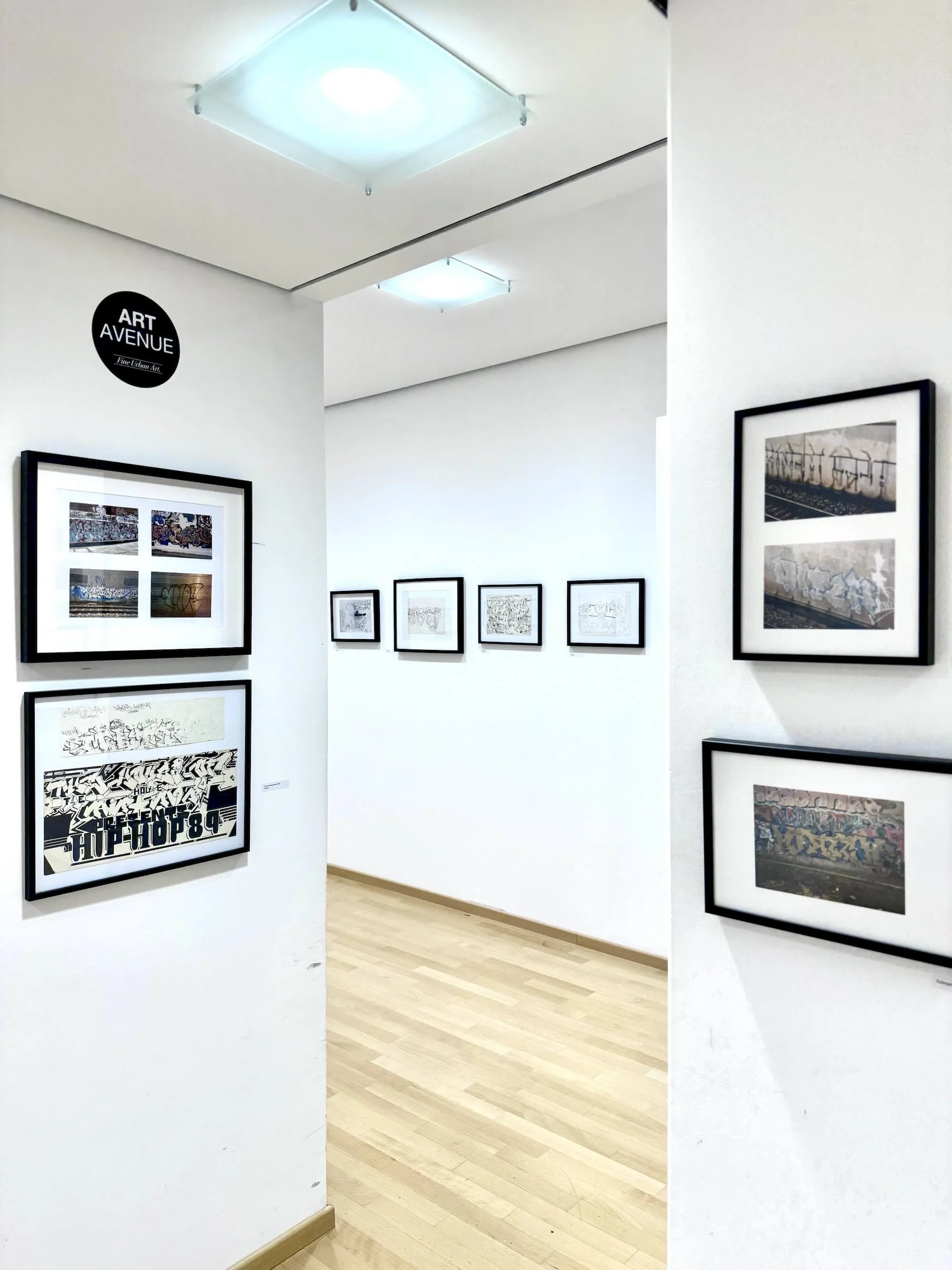 Galerie mit mehreren Kunstwerken in schwarzen Rahmen an weißen Wänden, Holzfußboden, Beleuchtung an Decke, Hinweisschild "ART AVENUE" oben links