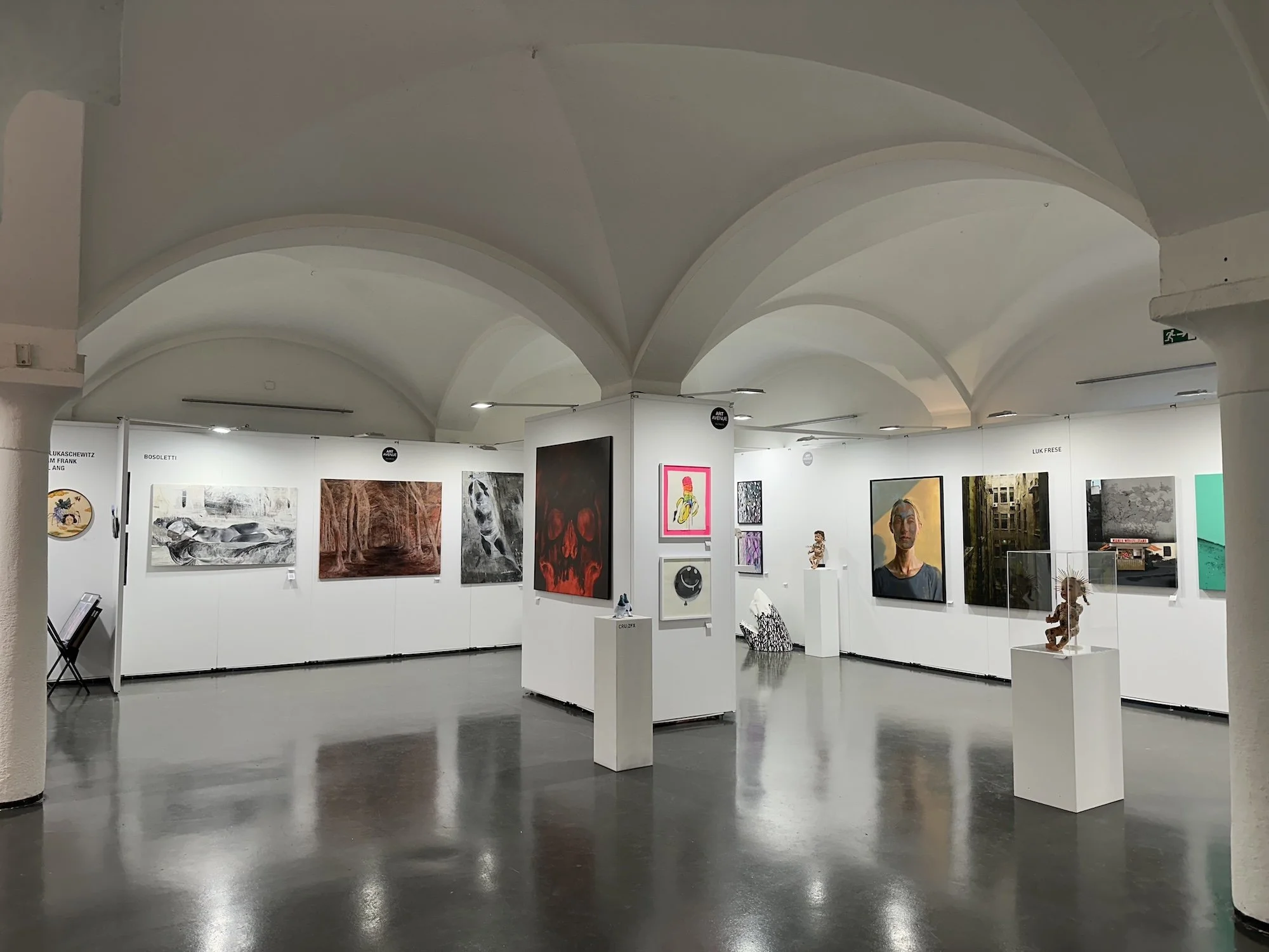 Ausstellung in einem Kunstmuseum mit Wandgemälden und Skulpturen, weiße Wände, helle Decken mit Bogen, reflektierende dunkle Boden, beleuchtete Kunstwerke auf Ständern und Wänden.